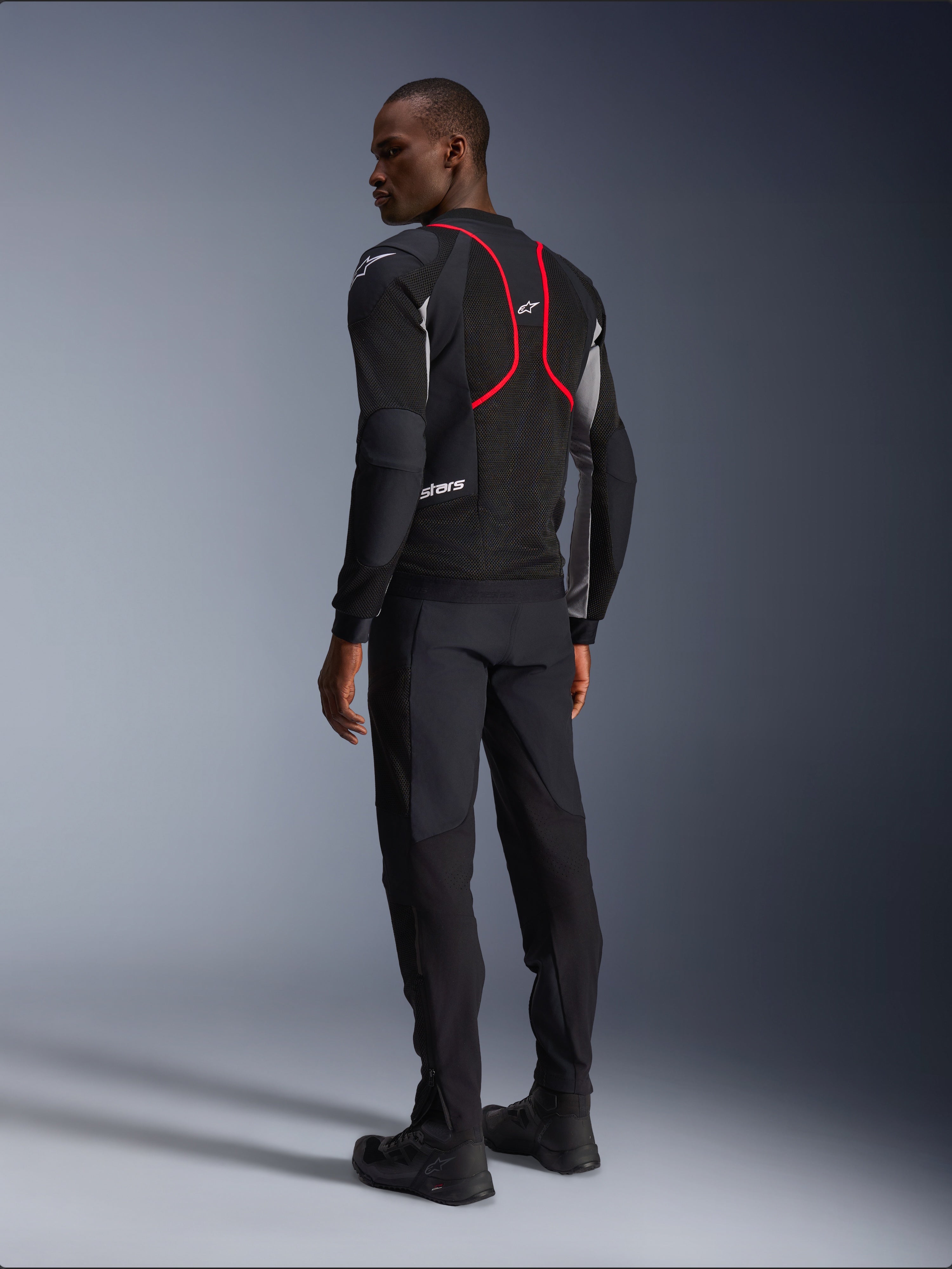 Alpinestars Troop-Air Pro Jacke, Motorradjacke, Schwarz, Eschegrau und Hellrot, Rückansicht am Modell, mit roten Akzentlinien und Mesh-Belüftungseinsätzen, getragen mit schwarzer Hose und Stiefeln, urbaner Motorradstil