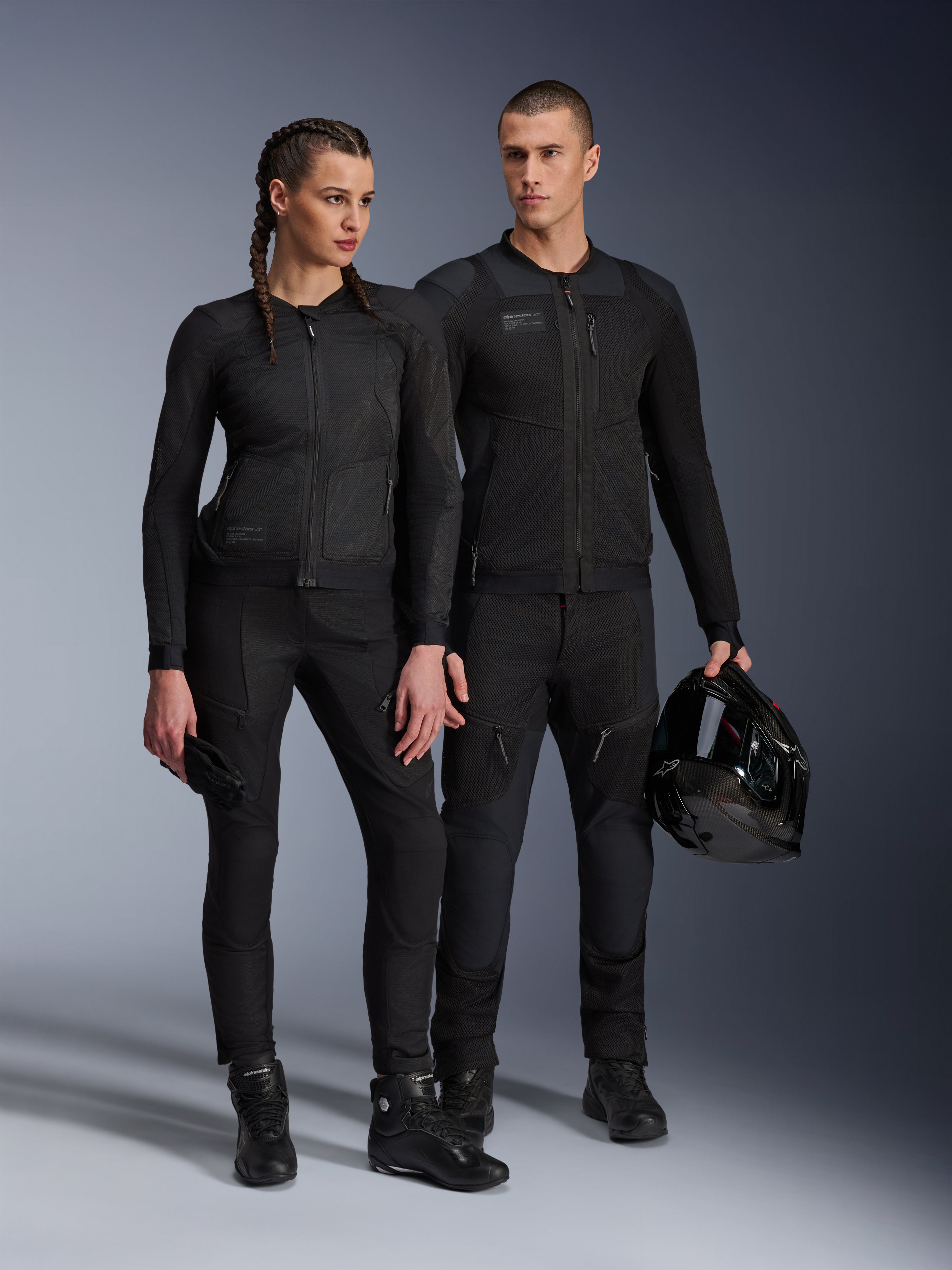 Alpinestars Stella Troop-Air Jacket, Man and woman wearing matching all-black technical motorcycle gear, featuring mesh construction for airflow, black pants and boots, man holding a black helmet, urban riding apparel.