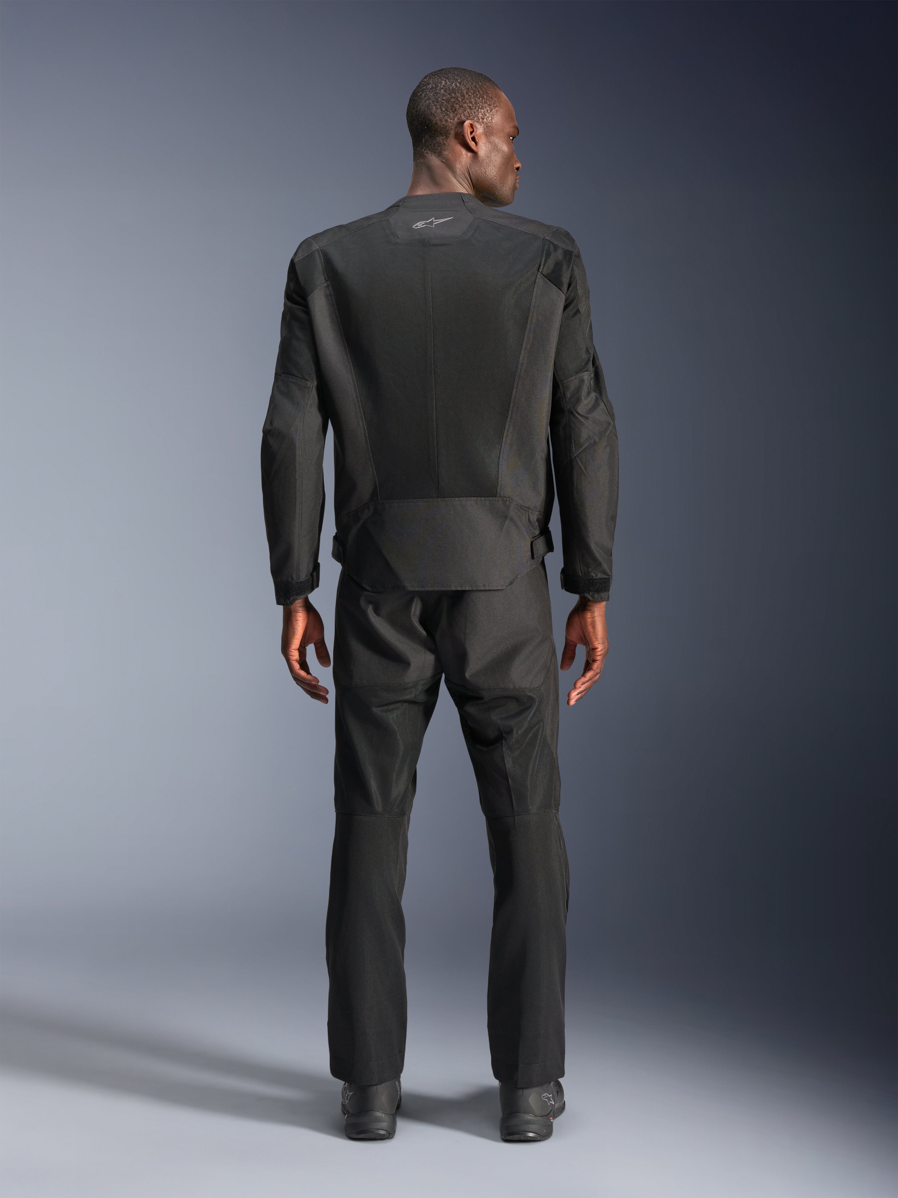 Rückansicht eines Mannes in der Alpinestars C-1 Air Jacke in Schwarz, urbane Motorradjacke mit Tech-Mesh-Einsätzen, passende schwarze Textilhose und Stiefel, schlichte Ästhetik mit reflektierendem Logo am oberen Rücken.