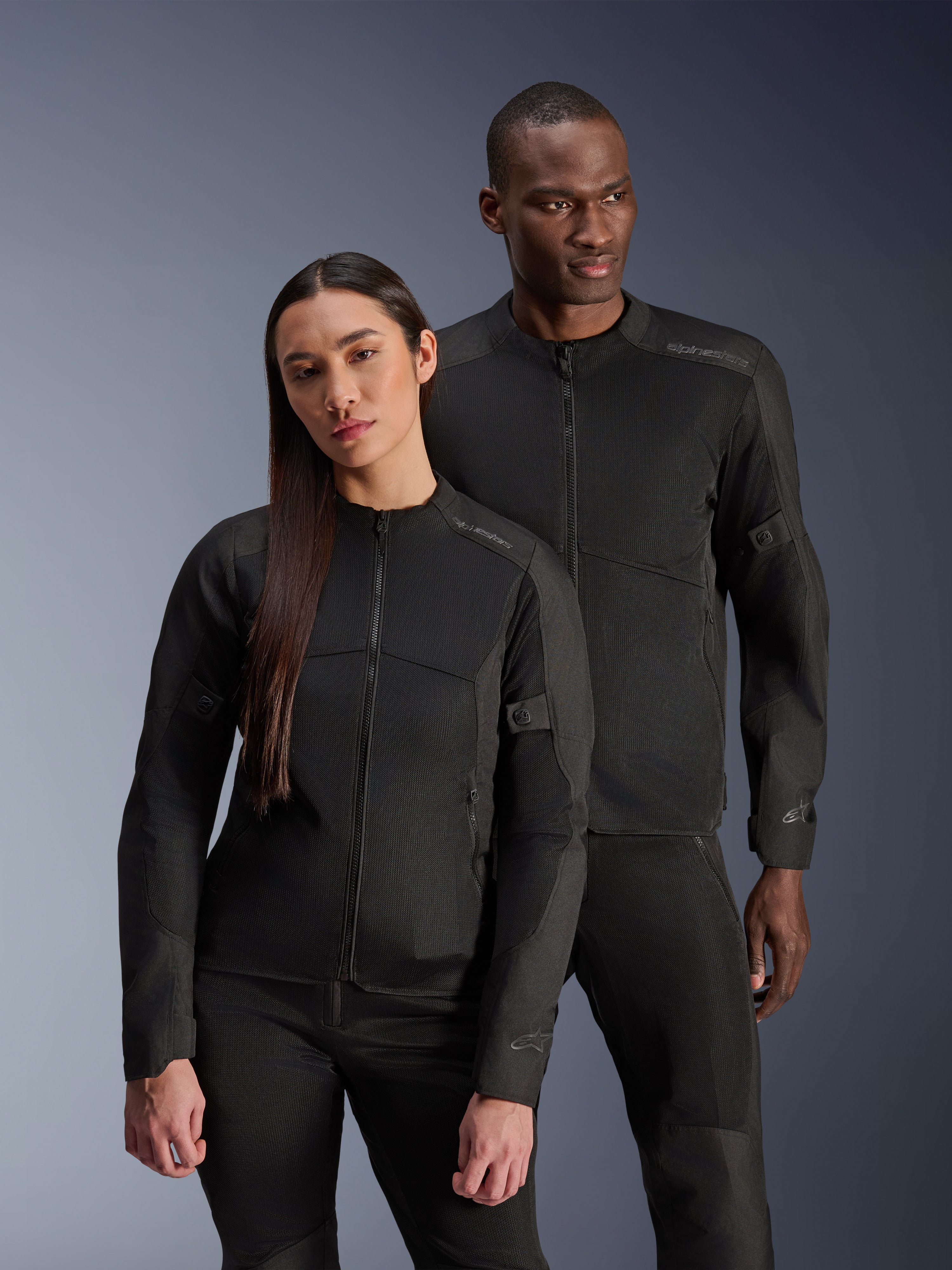 Ein Mann und eine Frau tragen die Alpinestars C-1 Air Jacke und die Stella C-1 Air Jacke, passende schwarze urbane Textil-Motorradjacken mit Mesh-Einsätzen zur Belüftung, kombiniert mit schwarzen Motorradhosen für einen klaren, modernen Look.