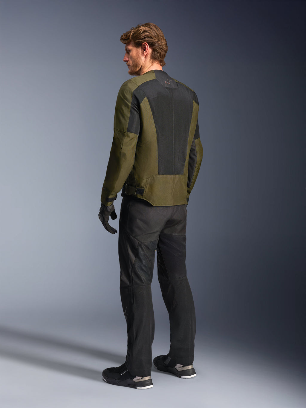 Männliches Model in Alpinestars C-1 Air Jacke in Khaki, urbane Motorradjacke, Rückansicht mit Fokus auf große schwarze Mesh-Paneele am Rücken, kombiniert mit schwarzer technischer Fahrhose und Handschuhen, moderne Motorradausrüstung für warmes Wetter