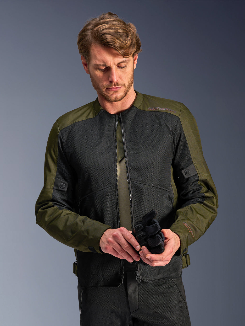 Mann in der Alpinestars C-1 Air Jacke in Khaki, urbane Motorradjacke mit olivgrünem Textil und schwarzen Mesh-Einsätzen, blickt nach unten und hält schwarze Motorradhandschuhe, cleaner und zeitgemäßer Stil für Straßenfahrten.
