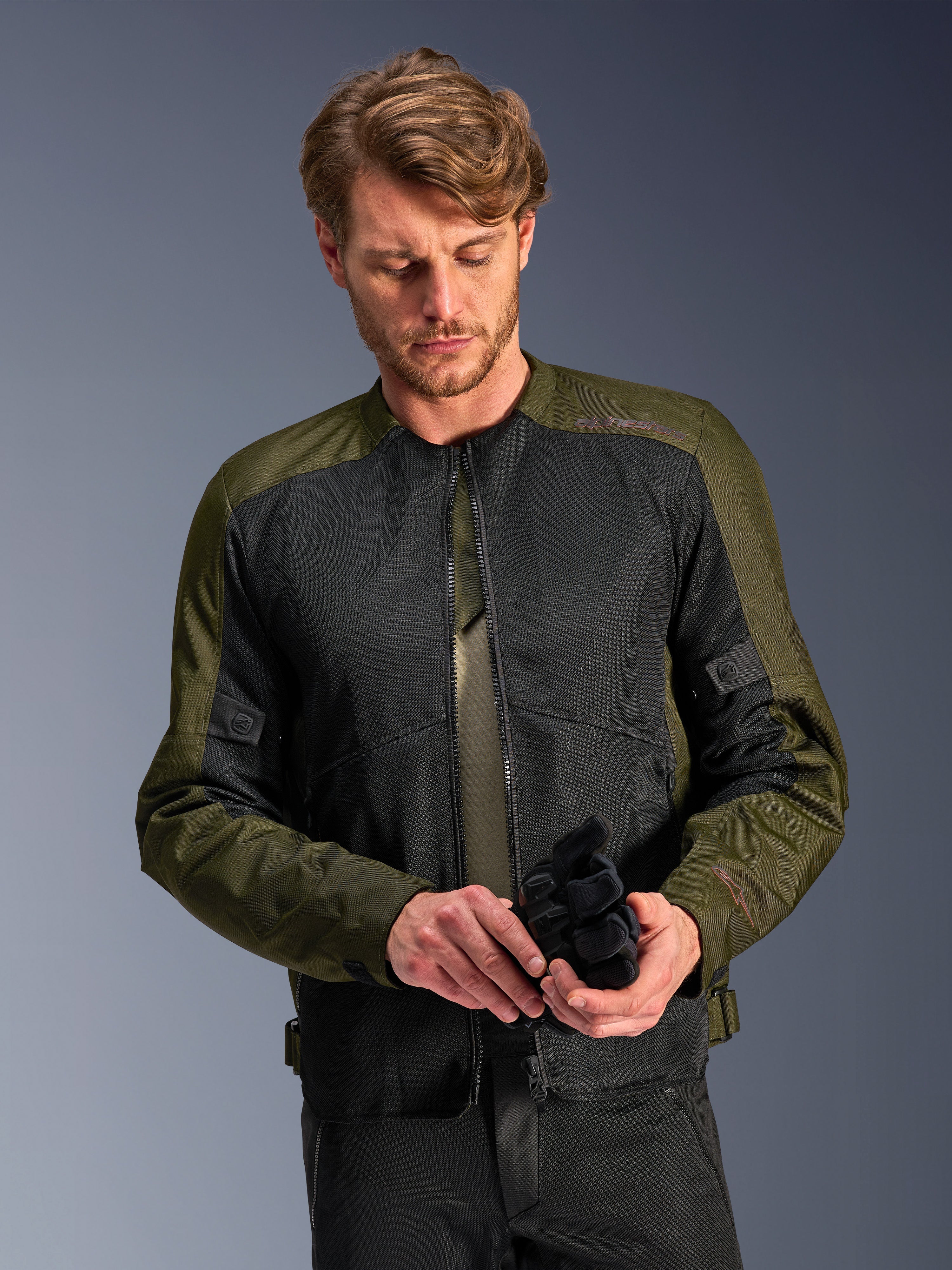Mann in der Alpinestars C-1 Air Jacke in Khaki, urbane Motorradjacke mit olivgrünem Textil und schwarzen Mesh-Einsätzen, blickt nach unten und hält schwarze Motorradhandschuhe, cleaner und zeitgemäßer Stil für Straßenfahrten.