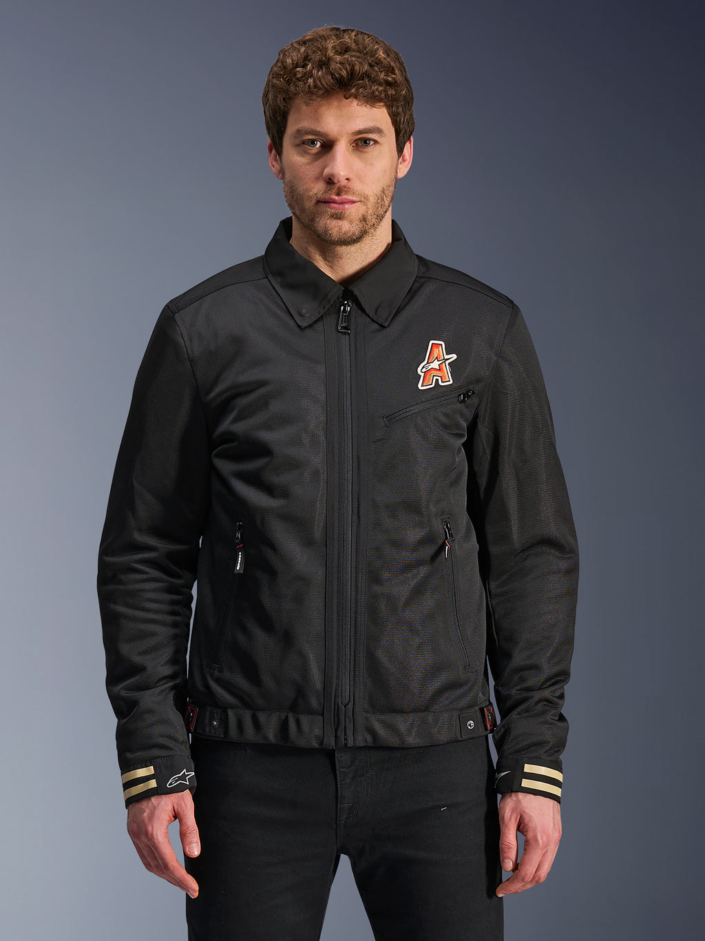 Alpinestars Ignite Air Jacket, Black motorcycle jacket, traditional shirt collar, orange and white 'A' logo on chest, beige striped cuffs with white star logo, urban contemporary riding gear