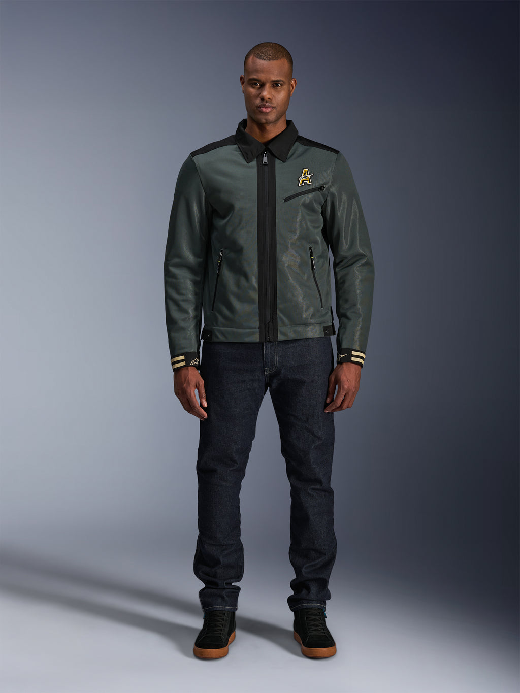 Alpinestars Ignite Air Jacke in Urban Gray Black Blue, Motorradjacke im Harrington-Stil mit Hemdkragen und Mesh-Konstruktion, getragen von einem Mann mit dunklen Denim-Jeans und schwarzen Sneakern, urbane Streetwear für Fahrer.