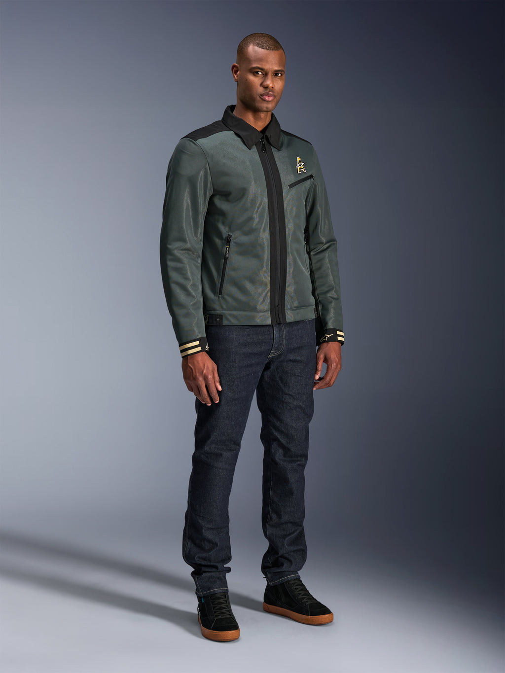 Alpinestars Ignite Air Jacke, Urban Gray Black Blue, urbane Motorradjacke, getragen von einem Model mit dunkler Denim-Jeans und schwarzen Sneakern, mit Mesh-Hauptteil, traditionellem Hemdkragen, Harrington-inspirierter Silhouette und goldenem Alpinestars-Branding.