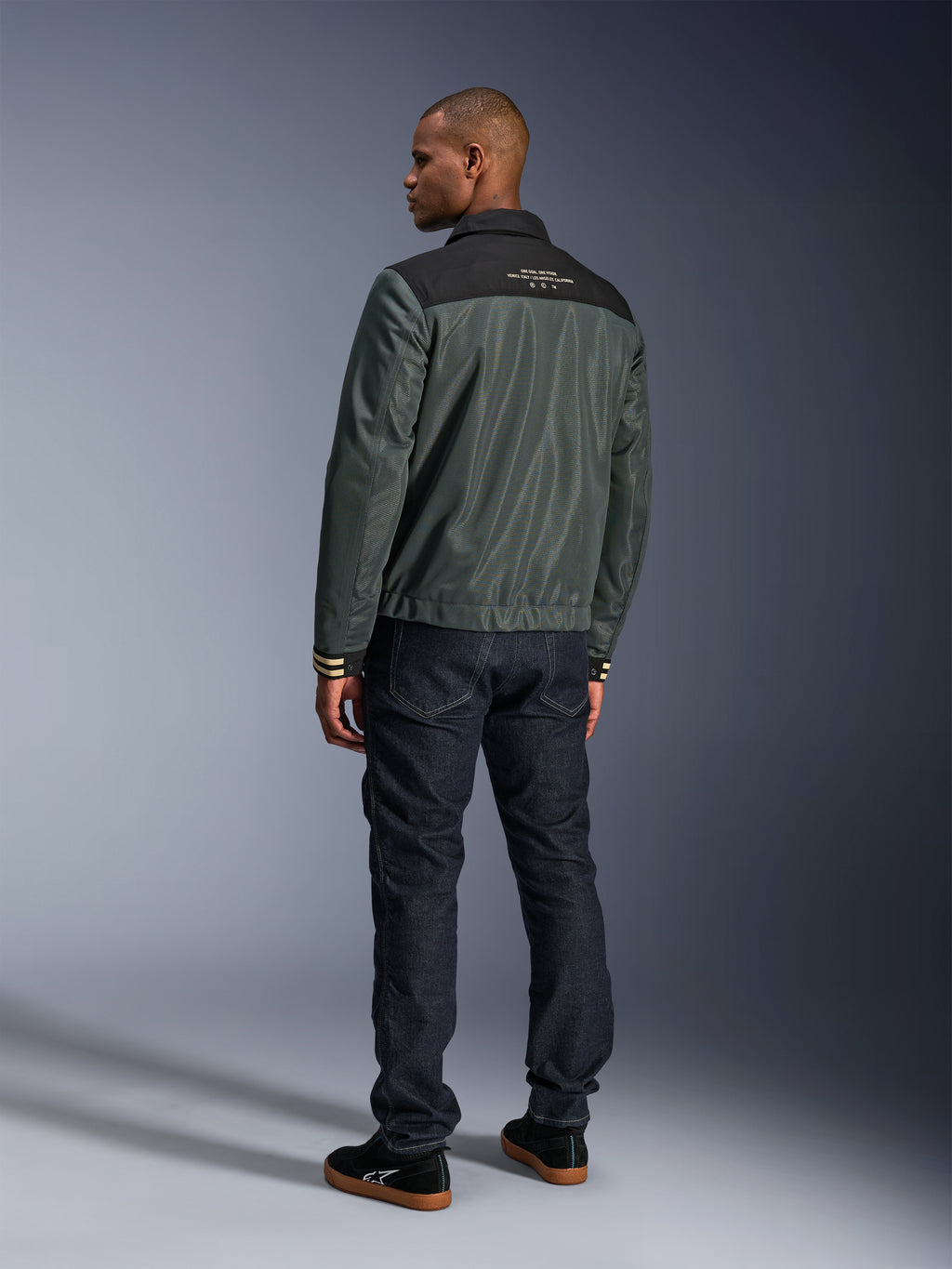 Alpinestars Ignite Air Jacke in Urban Gray Black Blue, Rückansicht am Modell, mit Mesh-Hauptteil, schwarzer Schulterpasse und weißem Text, gestreiften Bündchen, kombiniert mit dunklen Denim-Jeans und schwarzen Sneakern, urbane Lifestyle-Motorradbekleidung