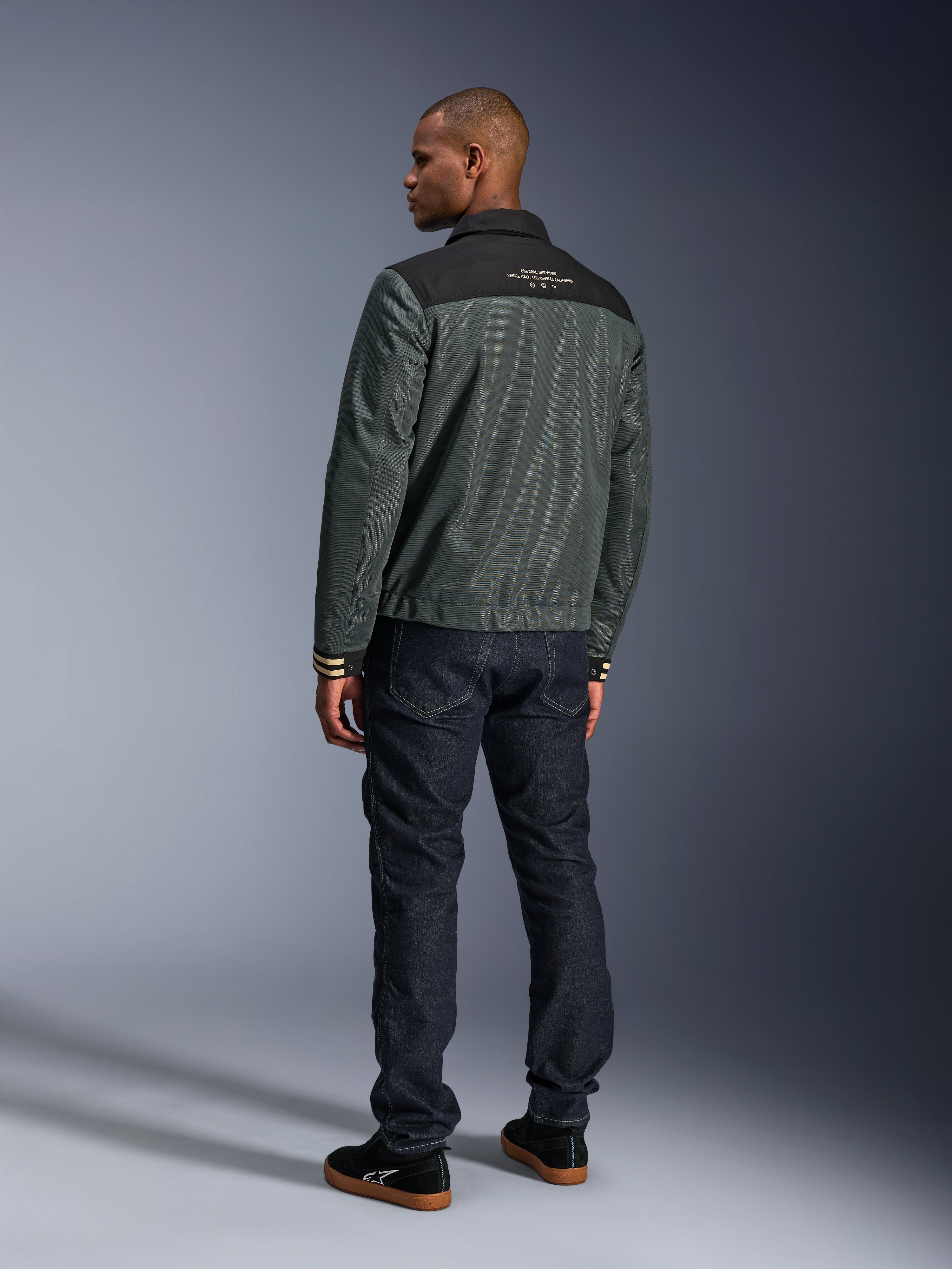 Alpinestars Ignite Air Jacke in Urban Gray Black Blue, Rückansicht am Modell, mit Mesh-Hauptteil, schwarzer Schulterpasse und weißem Text, gestreiften Bündchen, kombiniert mit dunklen Denim-Jeans und schwarzen Sneakern, urbane Lifestyle-Motorradbekleidung