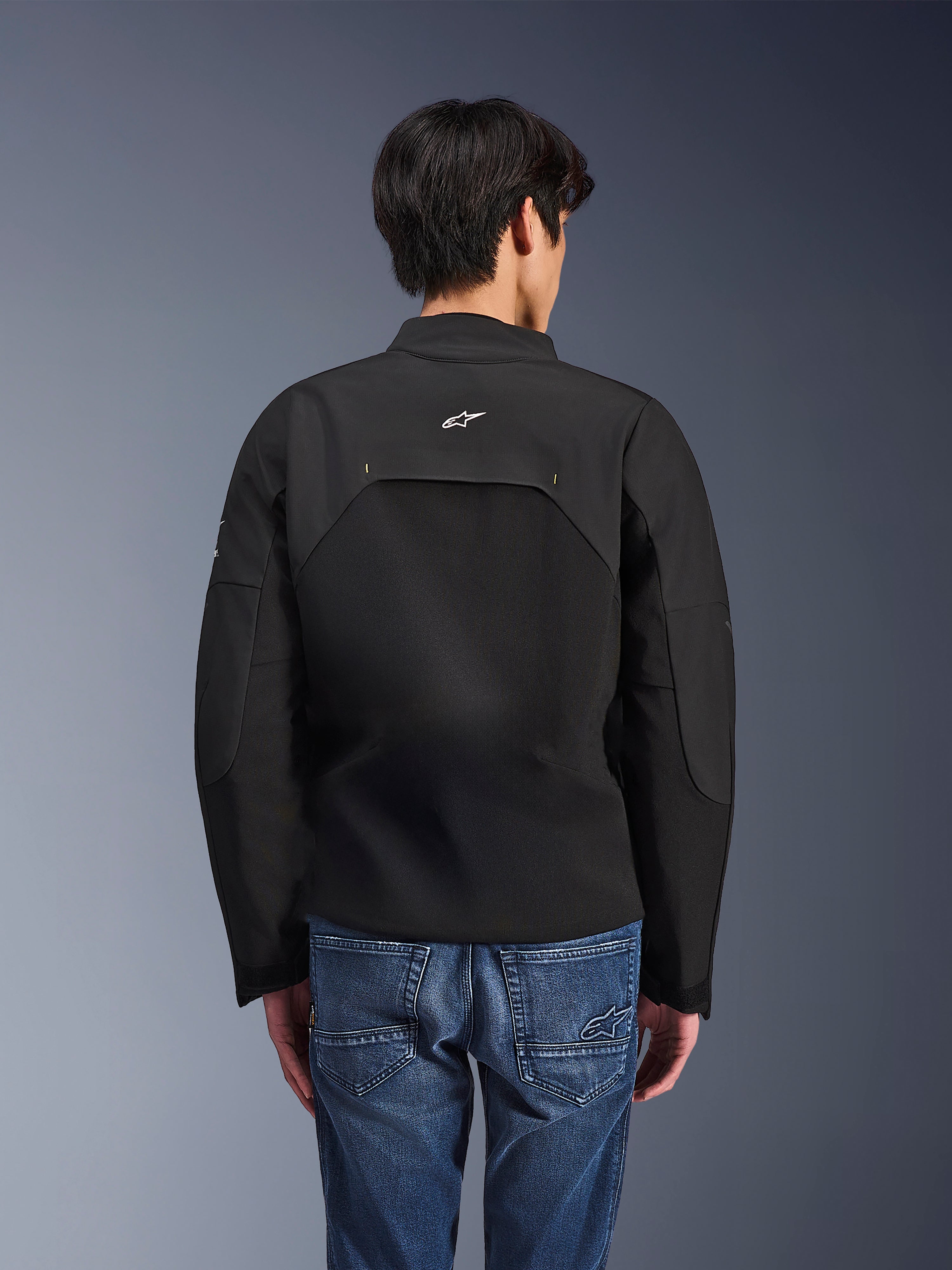 Alpinestars Provoke Jacke, Urbane Motorradjacke, Schwarz, Rückansicht mit weißem Astars-Logo und technischen Ripstop-Einsätzen, kombiniert mit blauen Denim-Jeans, modernes Design für den Stadtverkehr