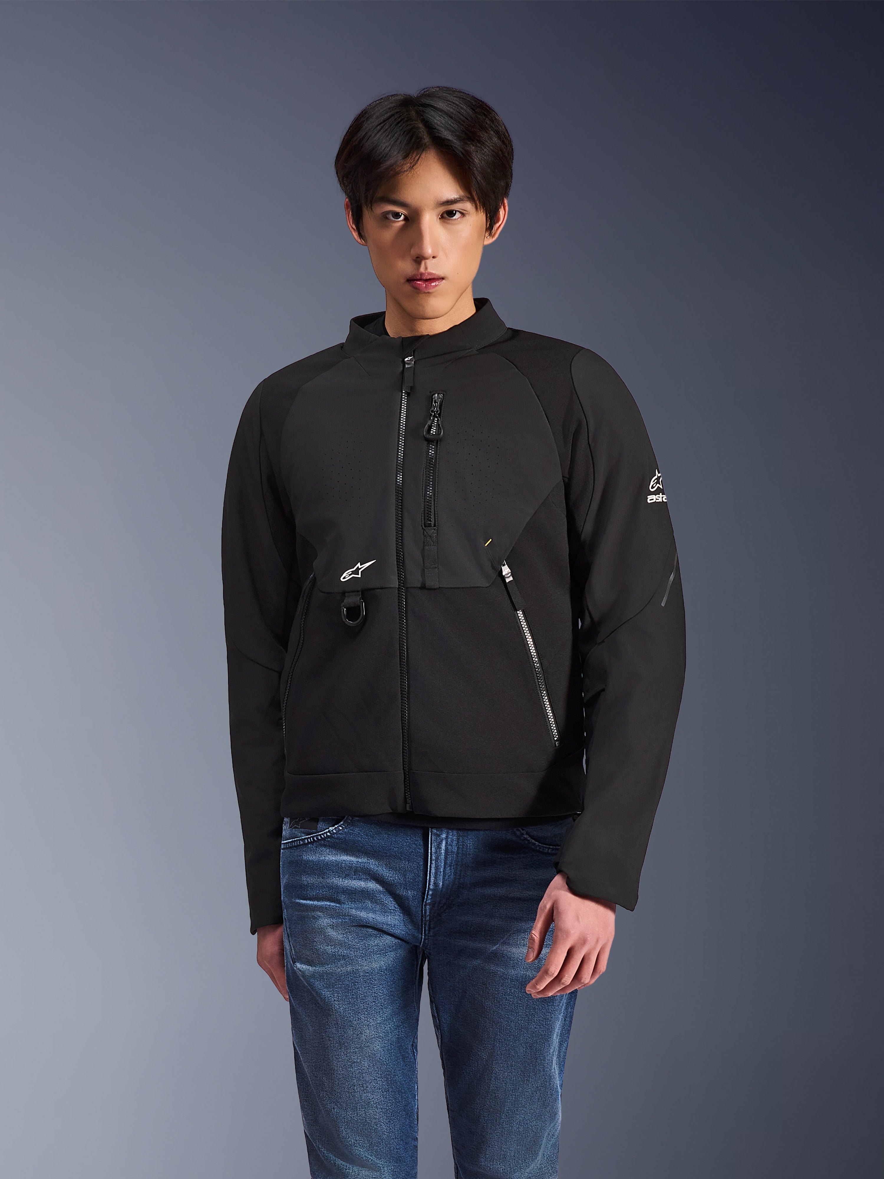 Alpinestars Provoke Jacke, schwarze Textil-Motorradjacke für den urbanen Einsatz, getragen von einem männlichen Model mit blauen Jeans, mit laserperforierten Brusteinsätzen, mehreren Reißverschlusstaschen, weißem A-star-Logo auf Brust und Ärmel, schlankes modernes Design für die Straße