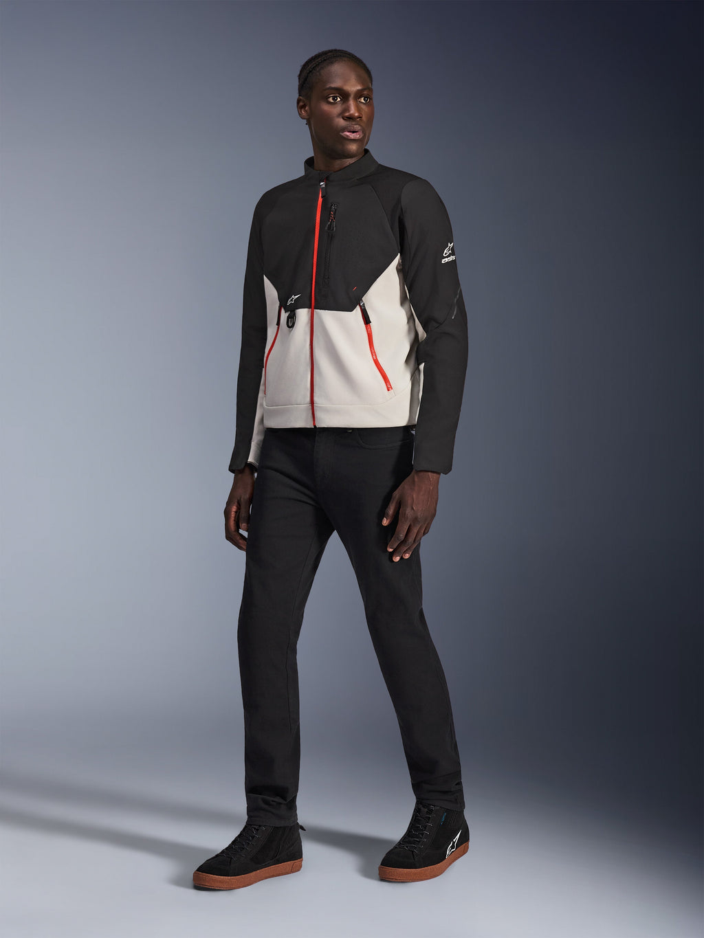 Modell trägt die Alpinestars Provoke Jacke in Black Metal Gray, mit markanten roten Front- und Taschenreißverschlüssen, kombiniert mit einer schwarzen Hose und schwarzen High-Top-Sneakern mit Gummisohlen, urbane zeitgenössische Motorradausrüstung für einen straßentauglichen Look.