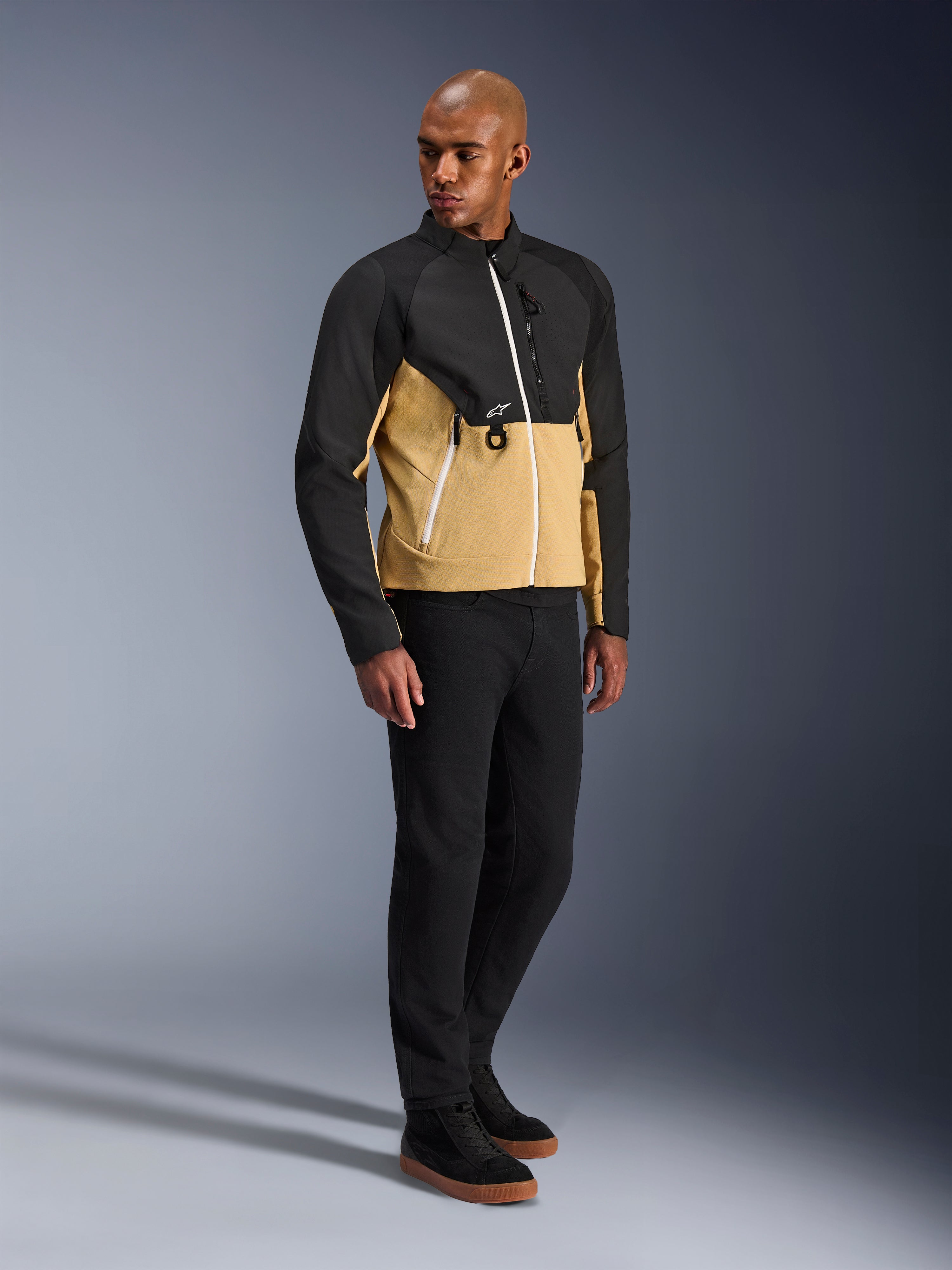 Alpinestars Provoke Jacke, urbane Motorradjacke, Black Camel, getragen von einem Model mit schwarzer Hose und Sneakern, zweifarbiges Textildesign mit schwarzem Ober- und camel-farbenem Unterkörper, weiße Reißverschlussdetails, moderne urbane Fahrerausrüstung