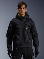 Alpinestars Mogress Air Jacke, urbane Motorradjacke, Schwarz mit Lime Punch-Akzenten, getragen von einem Model mit schwarzer Motorradhose und Handschuhen, Utility-inspiriertes Design mit Kapuze und weißem Stern-Logo auf der Brust, leichte Textilausrüstung für den Stadtverkehr