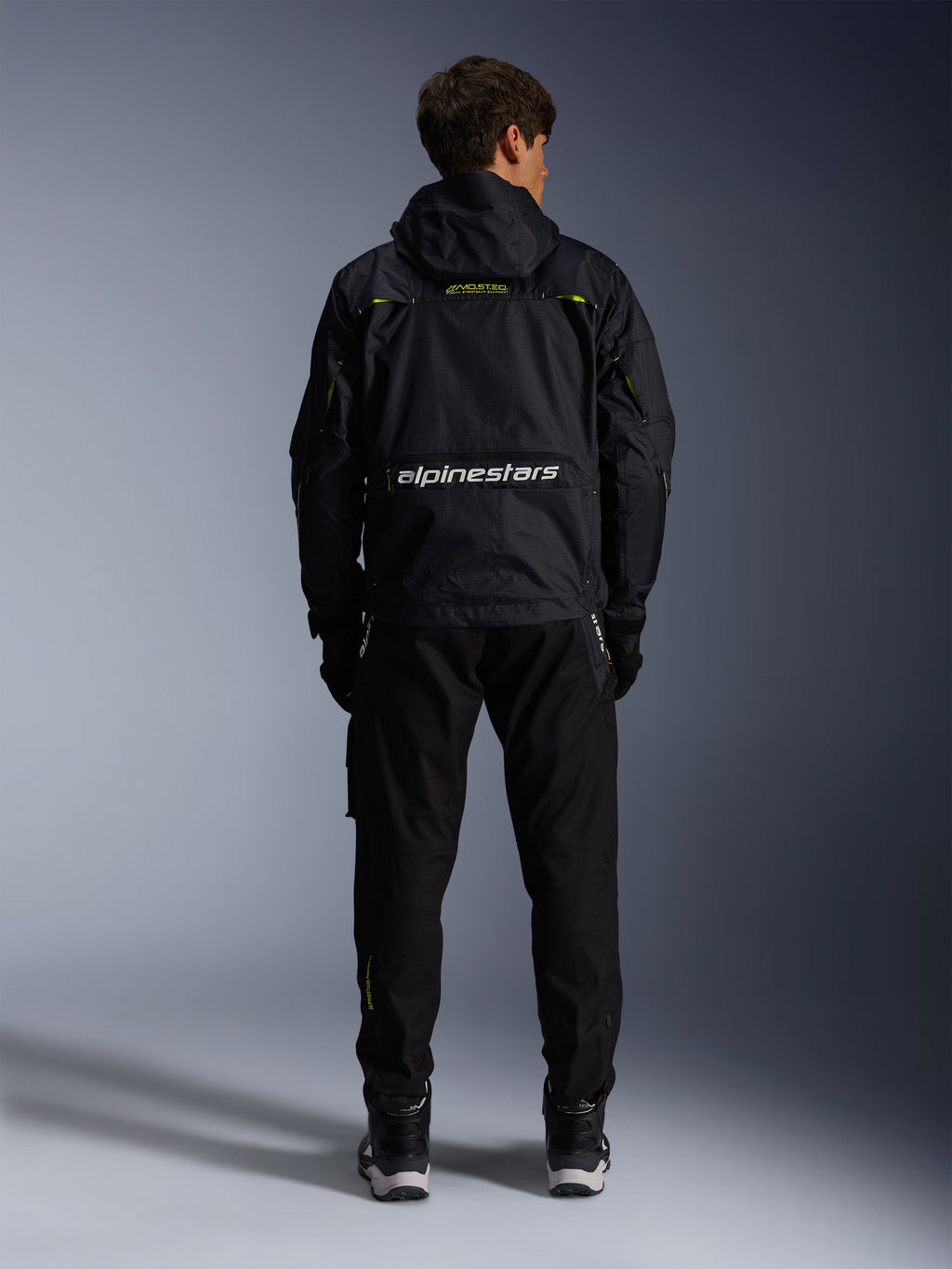 Alpinestars Mogress Air Jacke, urbane Motorradjacke, Rückansicht in Schwarz mit Lime Punch und weißen Akzenten, mit Alpinestars-Branding am unteren Rücken, getragen mit schwarzer Hose und Stiefeln.