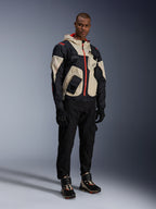 Model wearing the Alpinestars Mogress Air Jacket in Black, Almond, and Hot Orange, urban motorcycle jacket with a utility-inspired design, worn with black riding pants, black gloves, and coordinated technical boots, contemporary street-ready riding gear