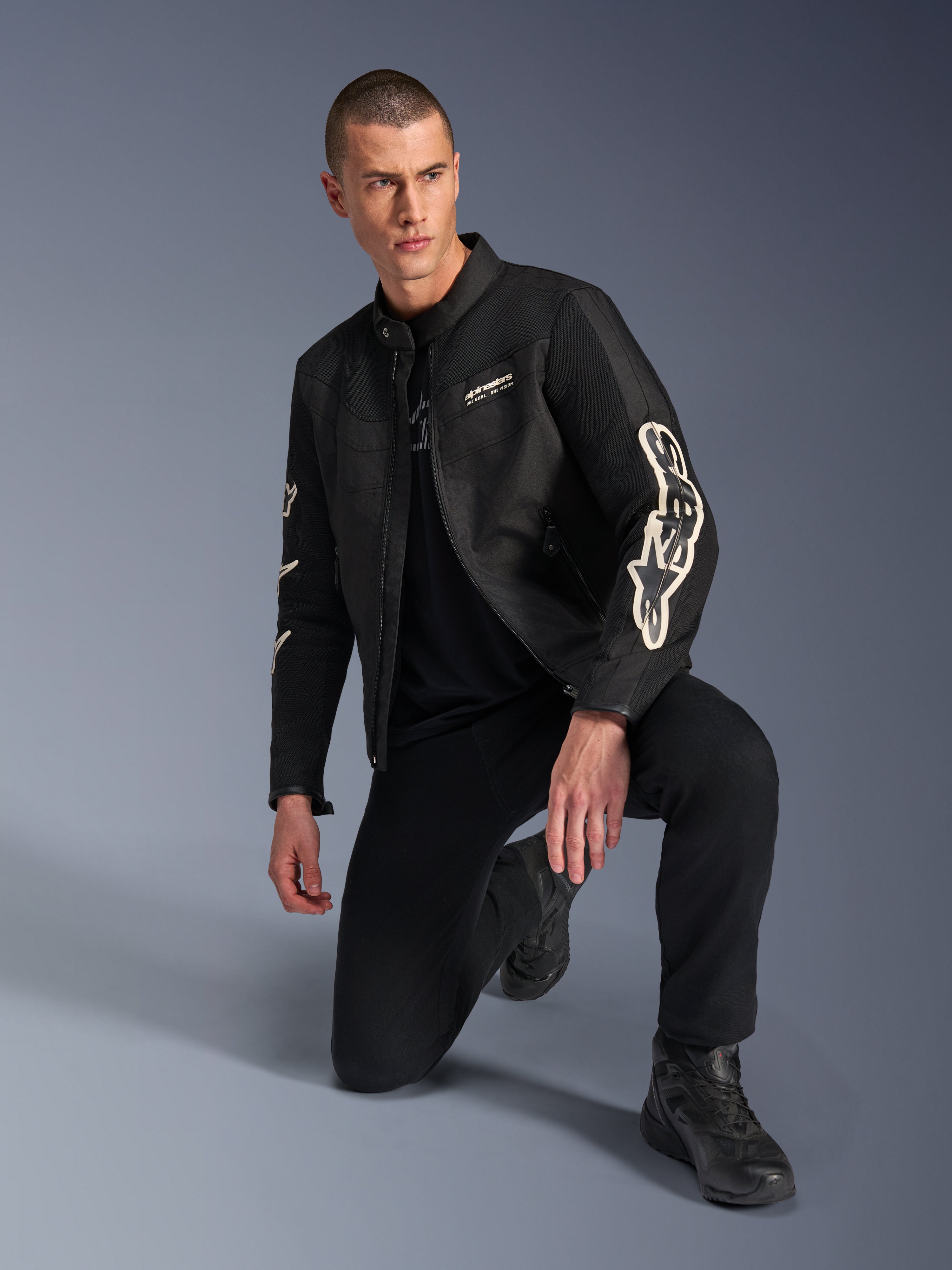 Male model wearing Alpinestars T-Dyno Air Jacket, Black textile motorcycle jacket with large white branding on sleeves, paired with black pants and riding boots, modern vintage-inspired design with 3D mesh panels.