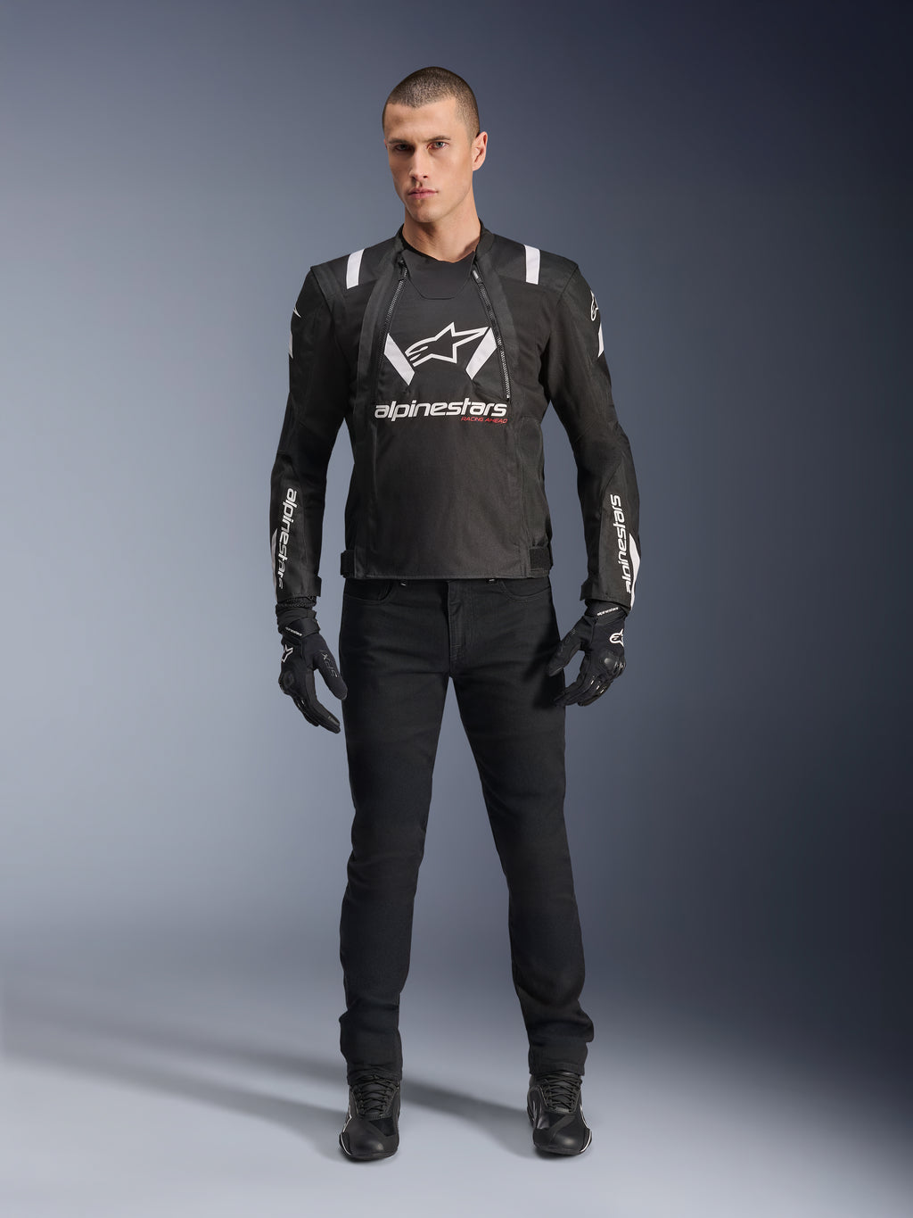 Alpinestars T-Stunt Air Jacke, schwarz-weiße Motorradjacke, getragen mit schwarzer Fahrhose, Handschuhen und Stiefeln, markantes weißes Alpinestars-Branding auf Brust und Ärmeln, sportliches Textildesign mit Mesh-Einsätzen zur Belüftung