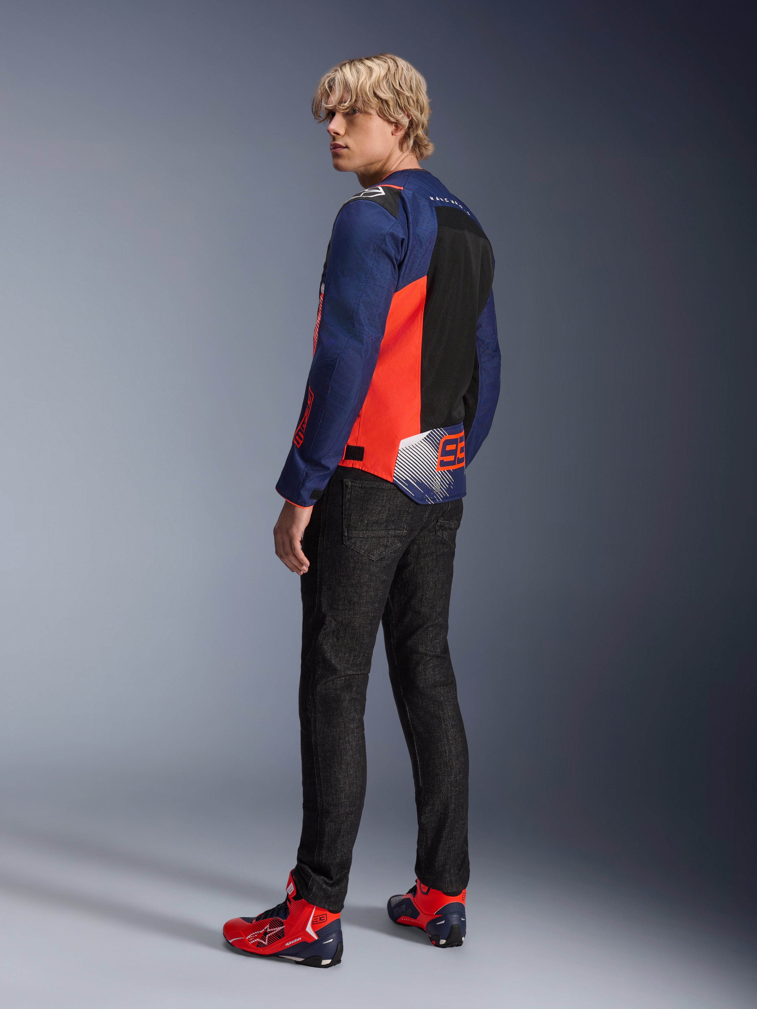 Alpinestars MM93 T-SPS Air V2 Jacke, Dunkelblau und Leuchtrot, von einem Modell von hinten getragen mit schwarzen Motorradjeans und passenden rot-blauen Schuhen, mit Mesh-Einsätzen und Marc Márquez 93 Branding.