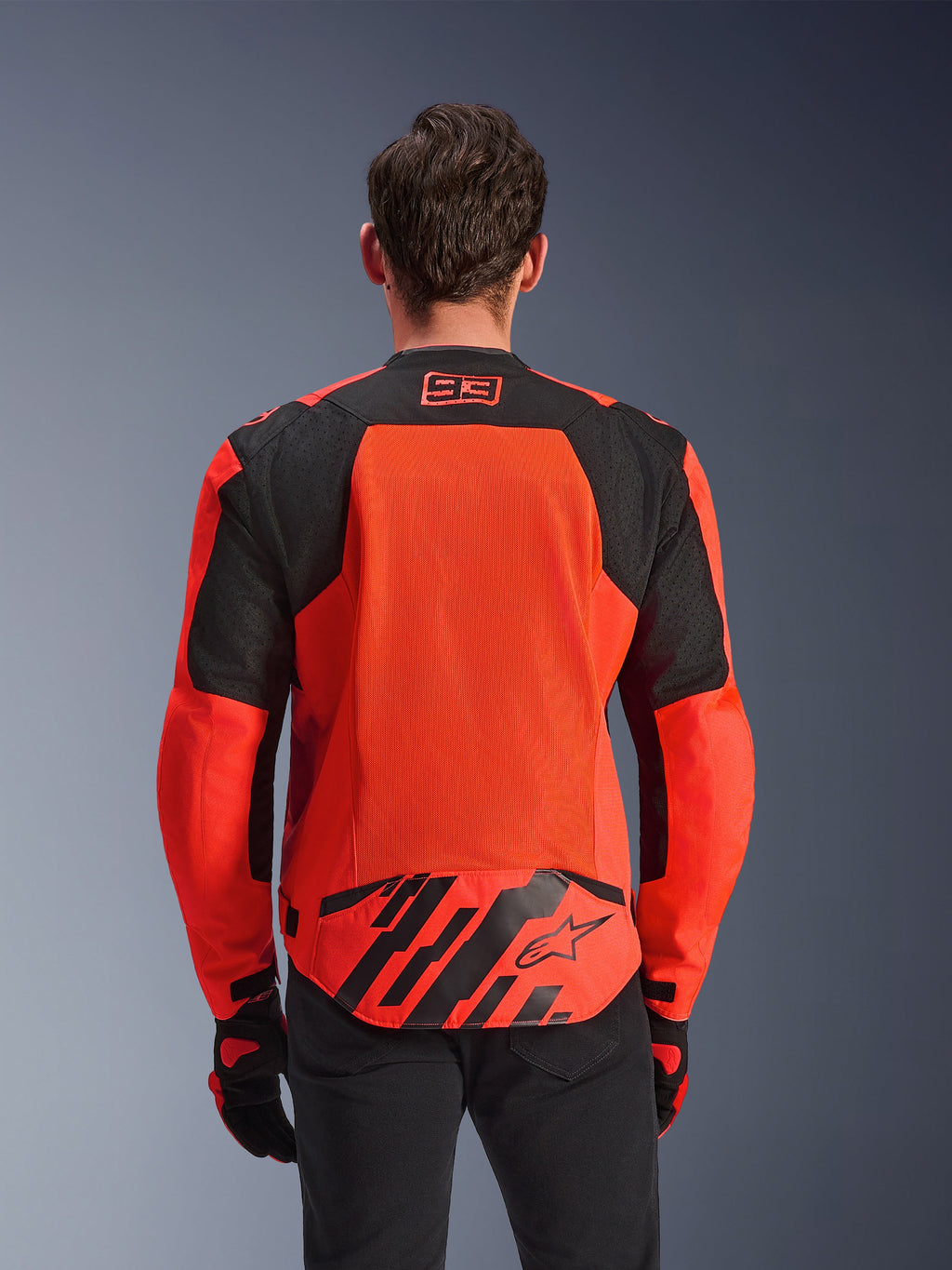 Alpinestars MM93 T-SPS Air V2 Jacket, Rear View, Red Fluo and Black, featuring 93 racing logo on the neck, large mesh panel on the back for airflow, lower back flap with Alpinestars star logo and geometric stripes, worn with black pants and riding gloves