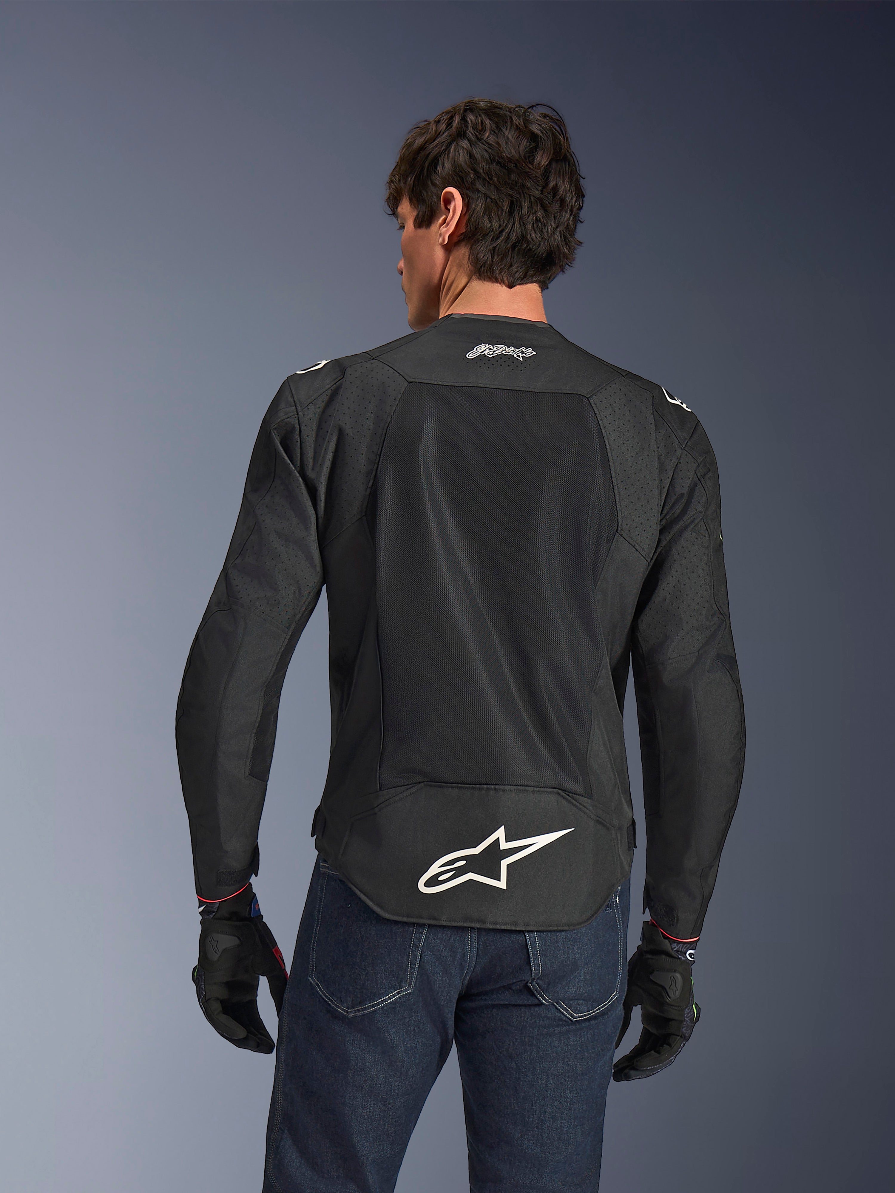 Alpinestars FQ20 T-SPS Air V2 Monster Jacke, Schwarz, Rückansicht mit großen Mesh-Belüftungseinsätzen und weißem Alpinestars-Logo am unteren Rücken, getragen mit blauen Jeans und schwarzen Motorradhandschuhen, urbaner Sport-Fahrstil.