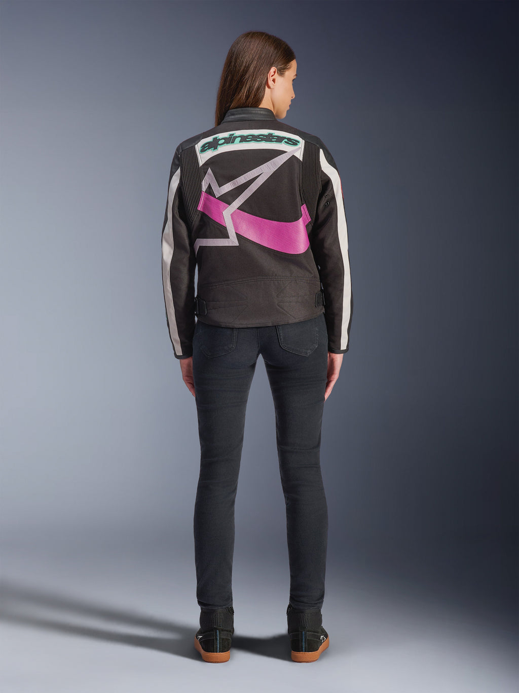 Rückansicht einer Frau in der Alpinestars Idles Jacke, Schwarz mit cremeweißen Streifen, mit einem großen lila-weißen Alpinestars Stern-Logo und grünem Schriftzug auf dem Rücken, kombiniert mit einer dunklen Slim-Fit-Jeans, vintage-inspirierte urbane Motorradbekleidung