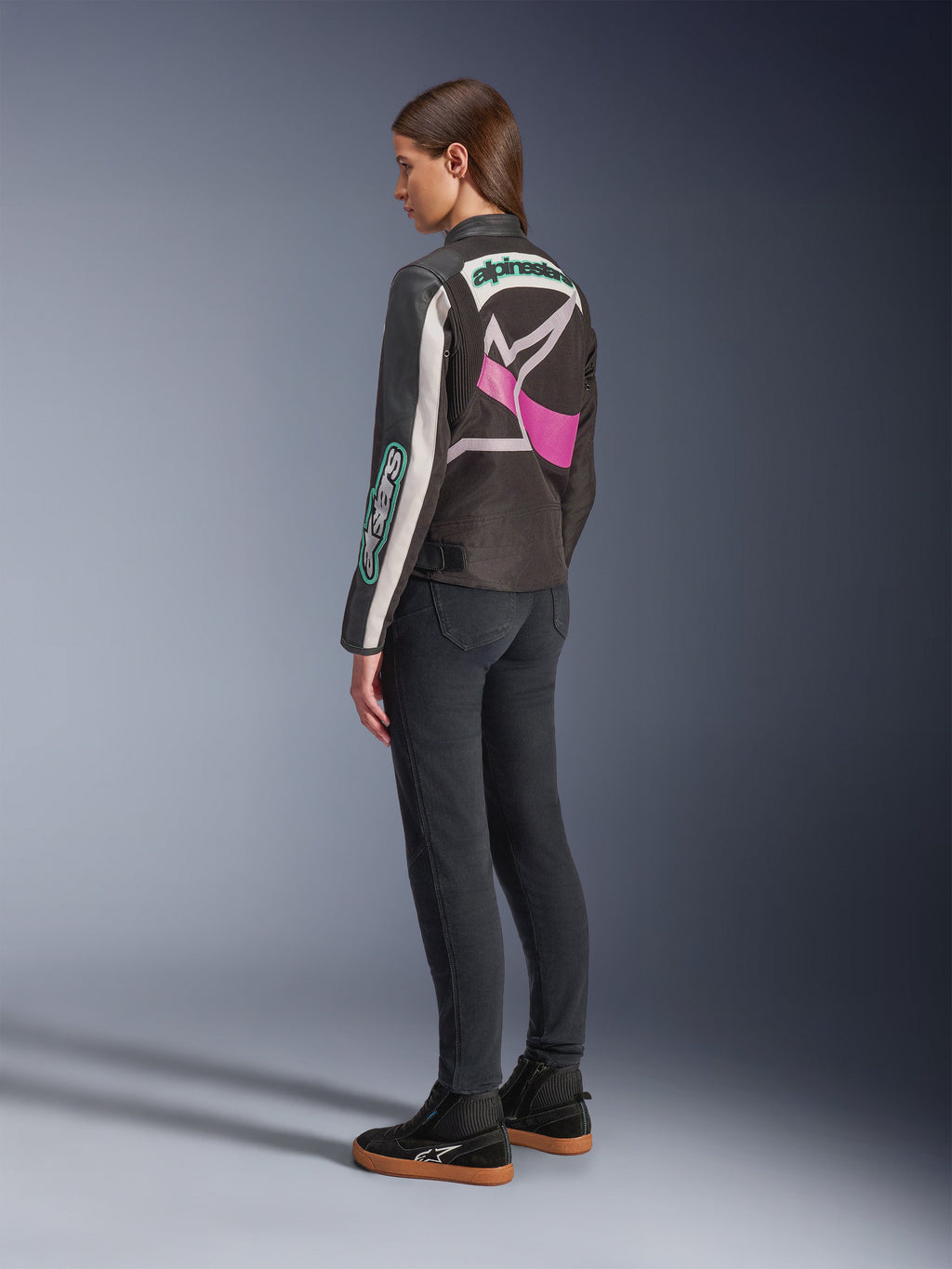 Alpinestars Idles Damenjacke, Motorradjacke, Schwarz, Off-White und Lila, Rückansicht am Modell, mit großem umlaufendem Alpinestars-Logo und von Vintage-Rennsport inspiriertem Design, kombiniert mit dunklen Jeans und schwarzen Schuhen.