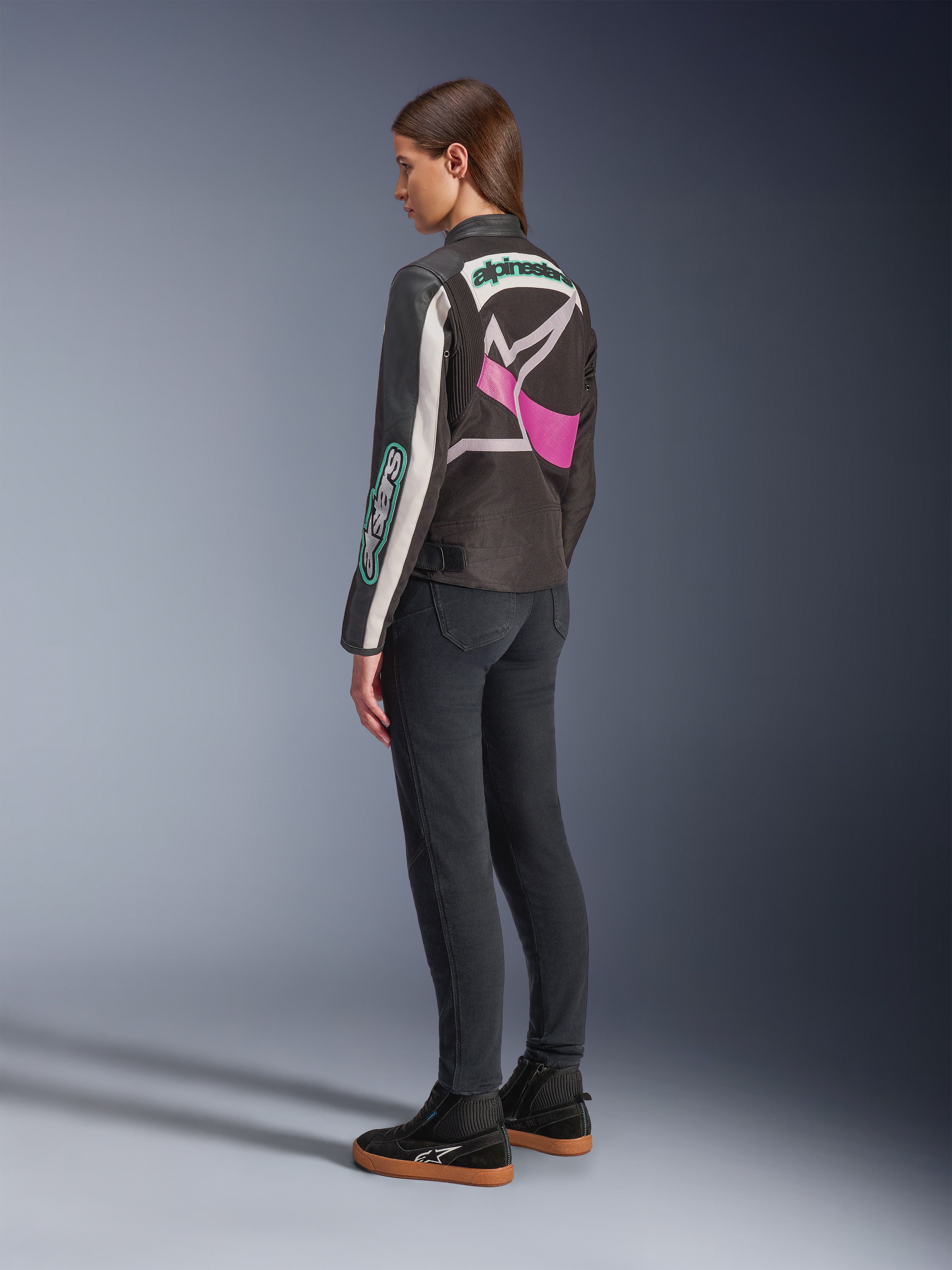 Alpinestars Idles Damenjacke, Motorradjacke, Schwarz, Off-White und Lila, Rückansicht am Modell, mit großem umlaufendem Alpinestars-Logo und von Vintage-Rennsport inspiriertem Design, kombiniert mit dunklen Jeans und schwarzen Schuhen.
