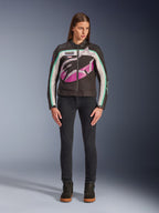 Frau in Alpinestars Idles Damenjacke, Schwarz, Off-White und Lila, von Vintage-Rennsport inspiriertes Design mit großem, umlaufendem Logo und türkisfarbenen Akzenten, kombiniert mit schwarzen Jeans und Motorrad-Sneakern, urbane Motorradbekleidung