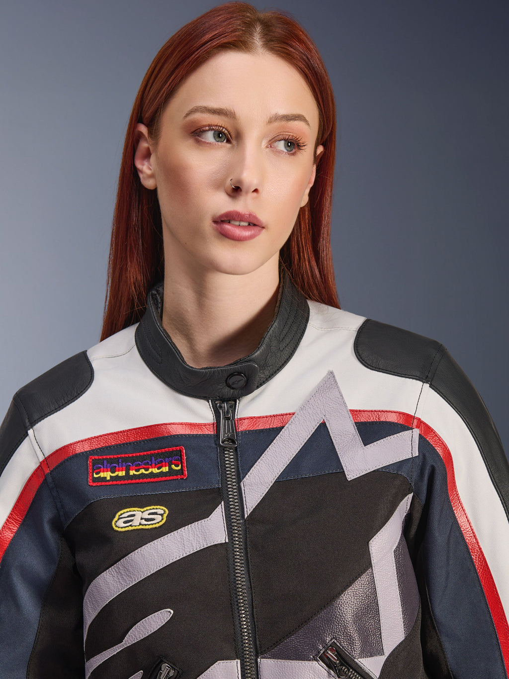 Alpinestars Idles Damenjacke, Nahaufnahme eines weiblichen Models mit roten Haaren, Farbkombination Off White, Flame Red und Schwarz, vintage-inspiriertes Renndesign mit Logo-Aufnähern und grauen geometrischen Sterngrafiken, urbane Motorradbekleidung.
