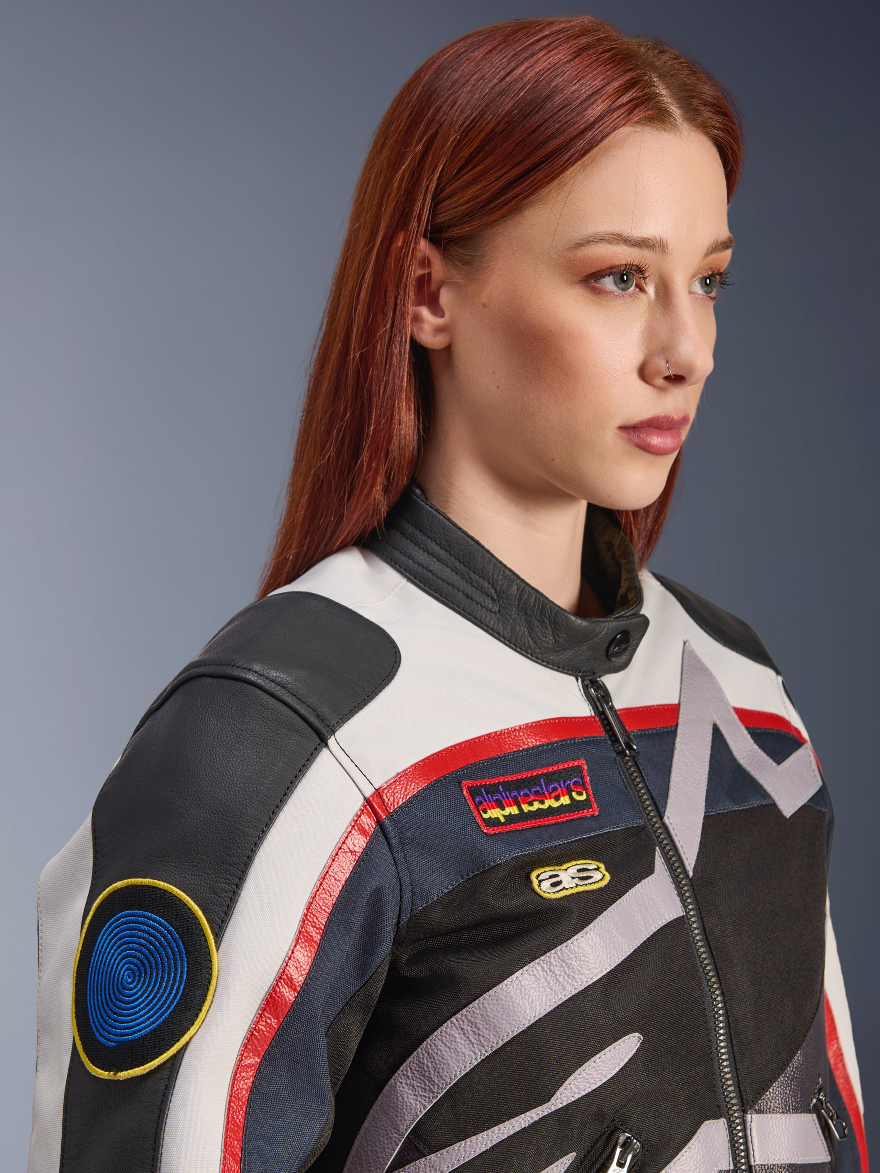Alpinestars Idles Damenjacke, Off-White, Flame-Red und Schwarz, mit von Vintage-Rennsport inspirierten Aufnähern und einem umlaufenden Logo, Textil- und Lederkonstruktion, urbaner Motorrad-Stil, getragen von einem Modell.