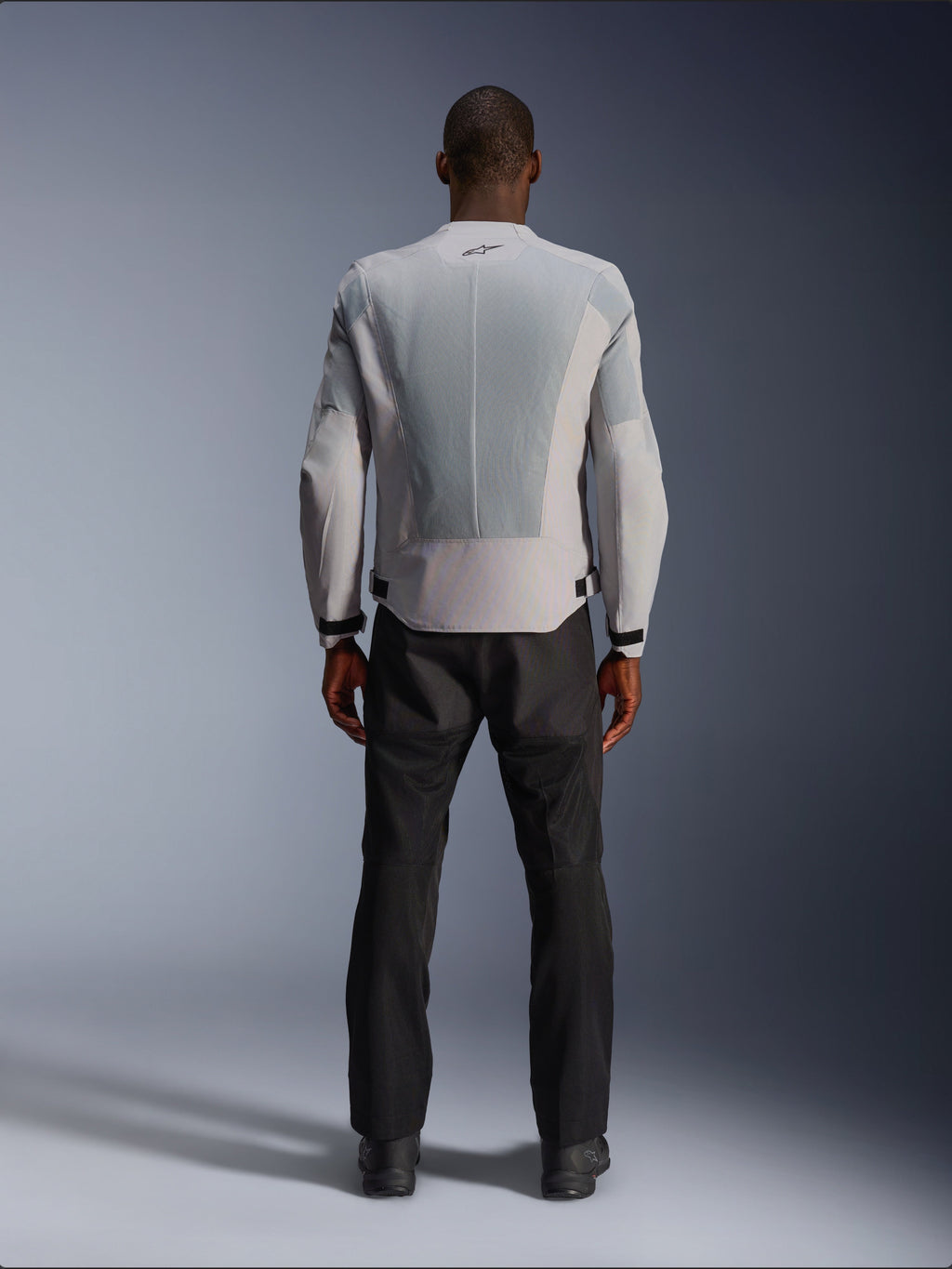 Alpinestars C-1 Air Jacke in Silber, Rückansicht an einem männlichen Model, Textil-Motorradjacke mit großen Tech-Mesh-Belüftungspanelen, kleinem schwarzen Logo im Nacken und Taillenverstellern, getragen mit einer schwarzen Motorradhose für einen modernen urbanen Look.
