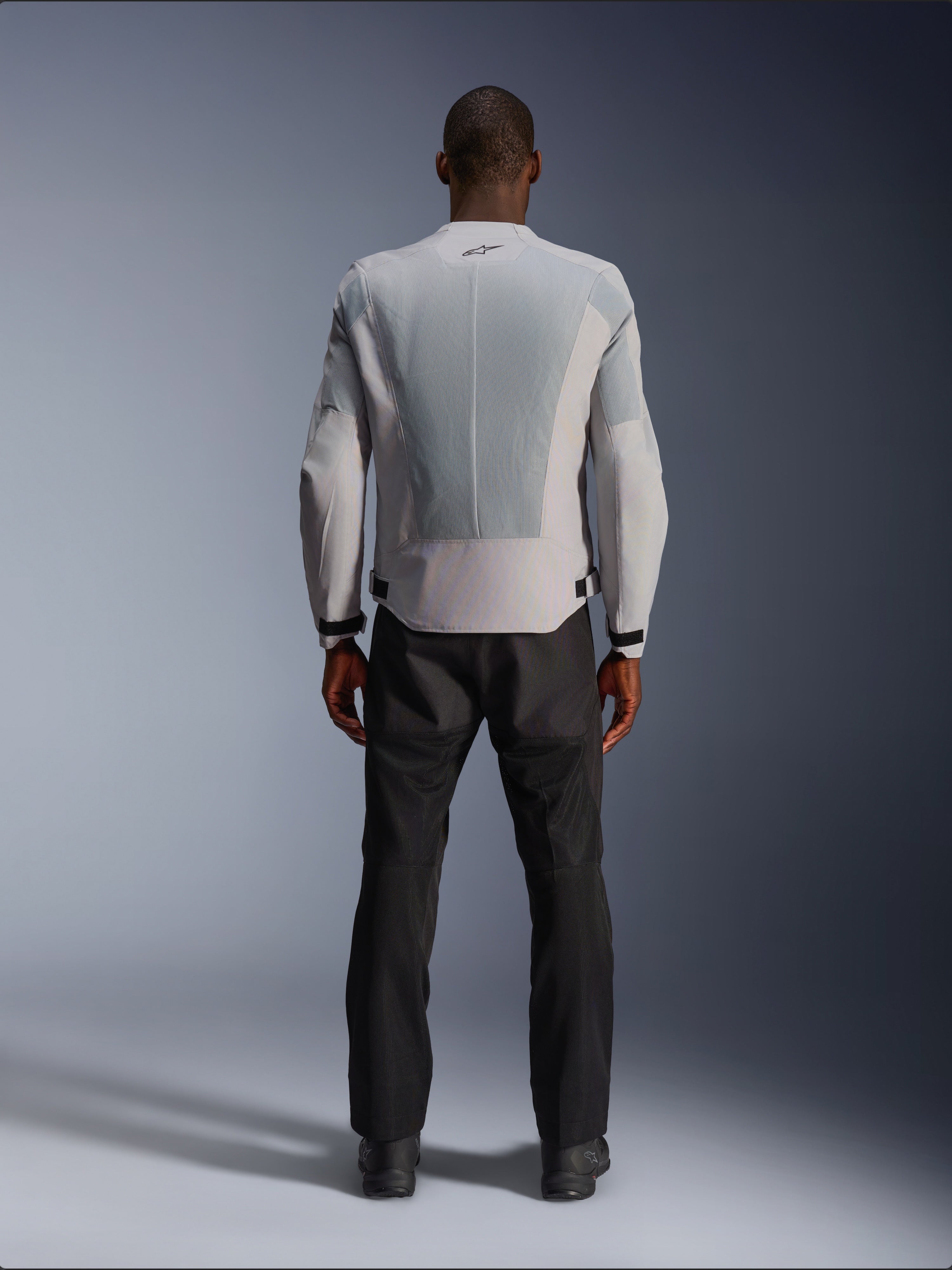 Alpinestars C-1 Air Jacke in Silber, Rückansicht an einem männlichen Model, Textil-Motorradjacke mit großen Tech-Mesh-Belüftungspanelen, kleinem schwarzen Logo im Nacken und Taillenverstellern, getragen mit einer schwarzen Motorradhose für einen modernen urbanen Look.