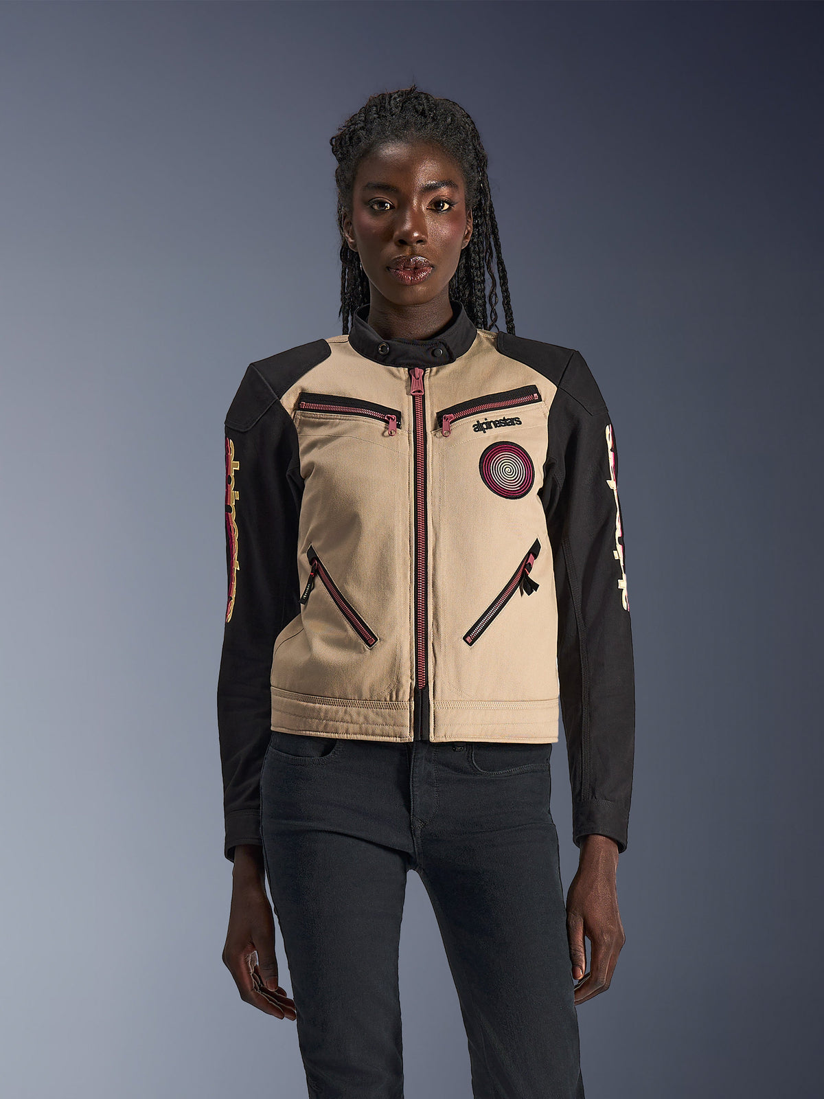 Alpinestars Stella Grace Jacke, Damen-Motorradjacke, Aluminum und Schwarz, mit einem von Workwear inspirierten Design, pink-roten Reißverschluss-Akzenten, einem spiralförmigen Brust-Aufnäher und langen schwarzen Ärmeln mit gelben und roten Grafiken, getragen von einem Model.