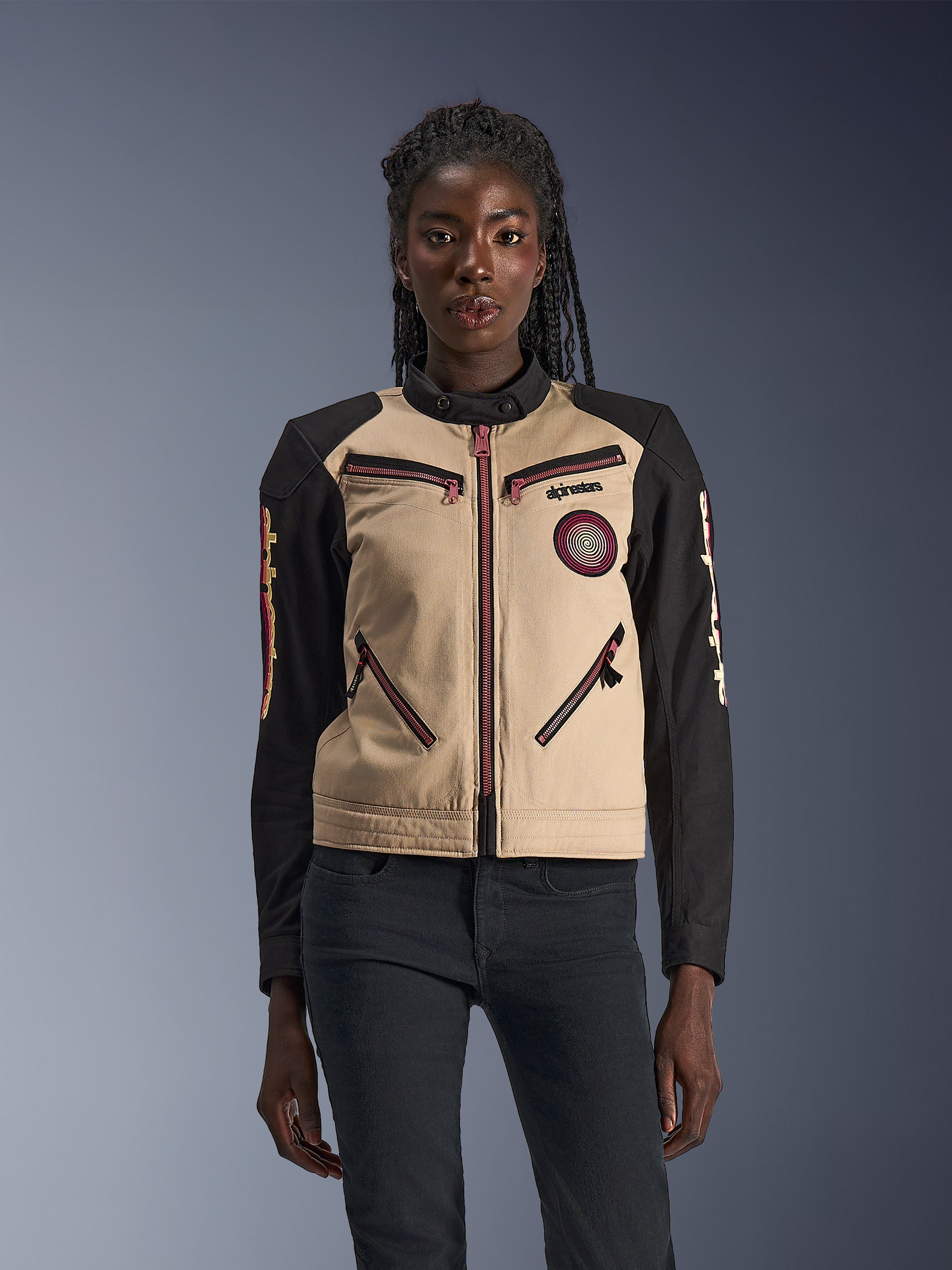 Alpinestars Stella Grace Jacket, Women's Motorcycle Jacket, Aluminum and Black, featuring a workwear-inspired design with pinkish-red zipper accents, a spiral chest patch, and long black sleeves with yellow and red graphics, worn by a model.