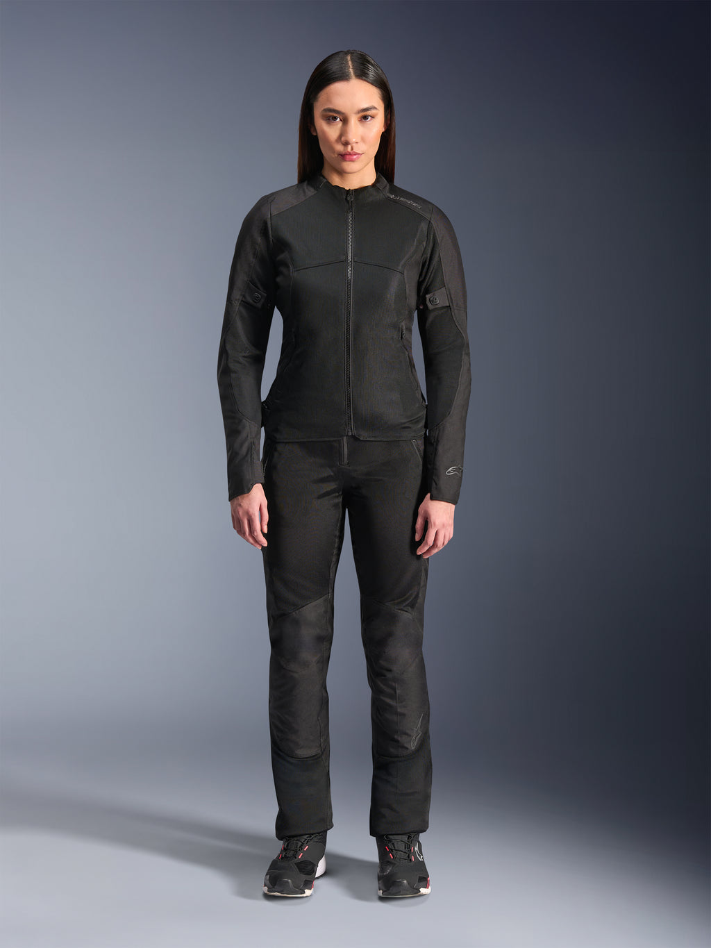 Frau trägt die Alpinestars Stella C-1 Air Jacke in Schwarz, kombiniert mit passender schwarzer Textil-Motorradhose und Motorradschuhen, mit einem eleganten urbanen Design und großen Mesh-Einsätzen für Luftzirkulation.