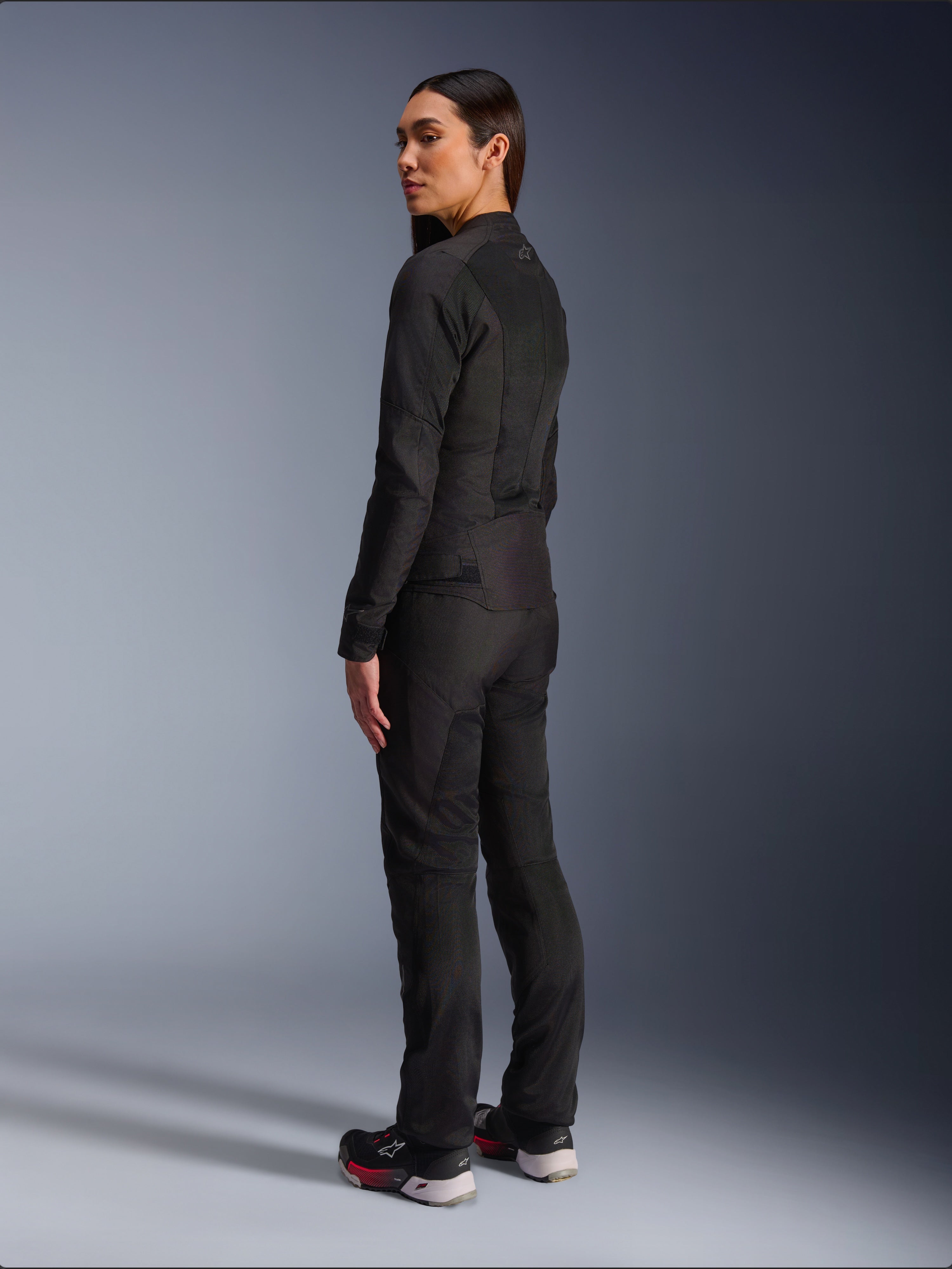 Frau in Alpinestars Stella C-1 Air Jacke in Schwarz, urbane Textil-Motorradjacke mit Mesh-Einsätzen, Rückansicht zeigt die schlanke Passform und das Logo am oberen Rücken, getragen mit passender schwarzer Hose und Sportschuhen, zeitgemäße Motorradausrüstung.