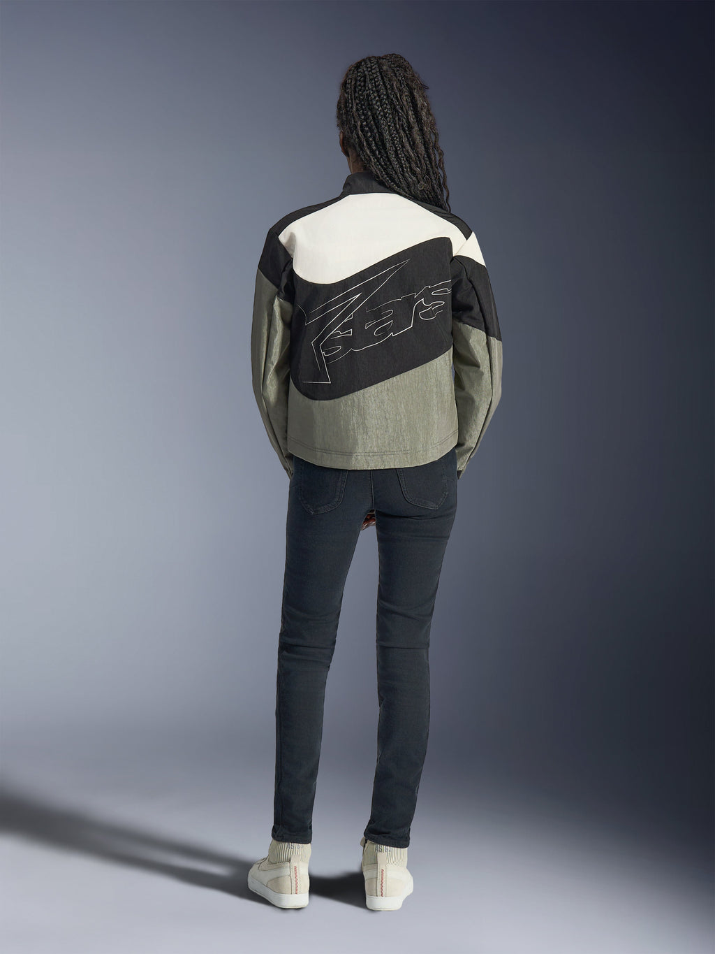 Rückansicht eines Models in der Alpinestars Stella Moshell Jacke in Schwarz, Grau und Off-White, mit asymmetrischem Design und großem, stilisiertem Logo auf dem Rücken, kombiniert mit einer dunklen Slim-Fit-Jeans und weißen Sneakern, urbane Streetwear-inspirierte Motorradbekleidung für Damen.