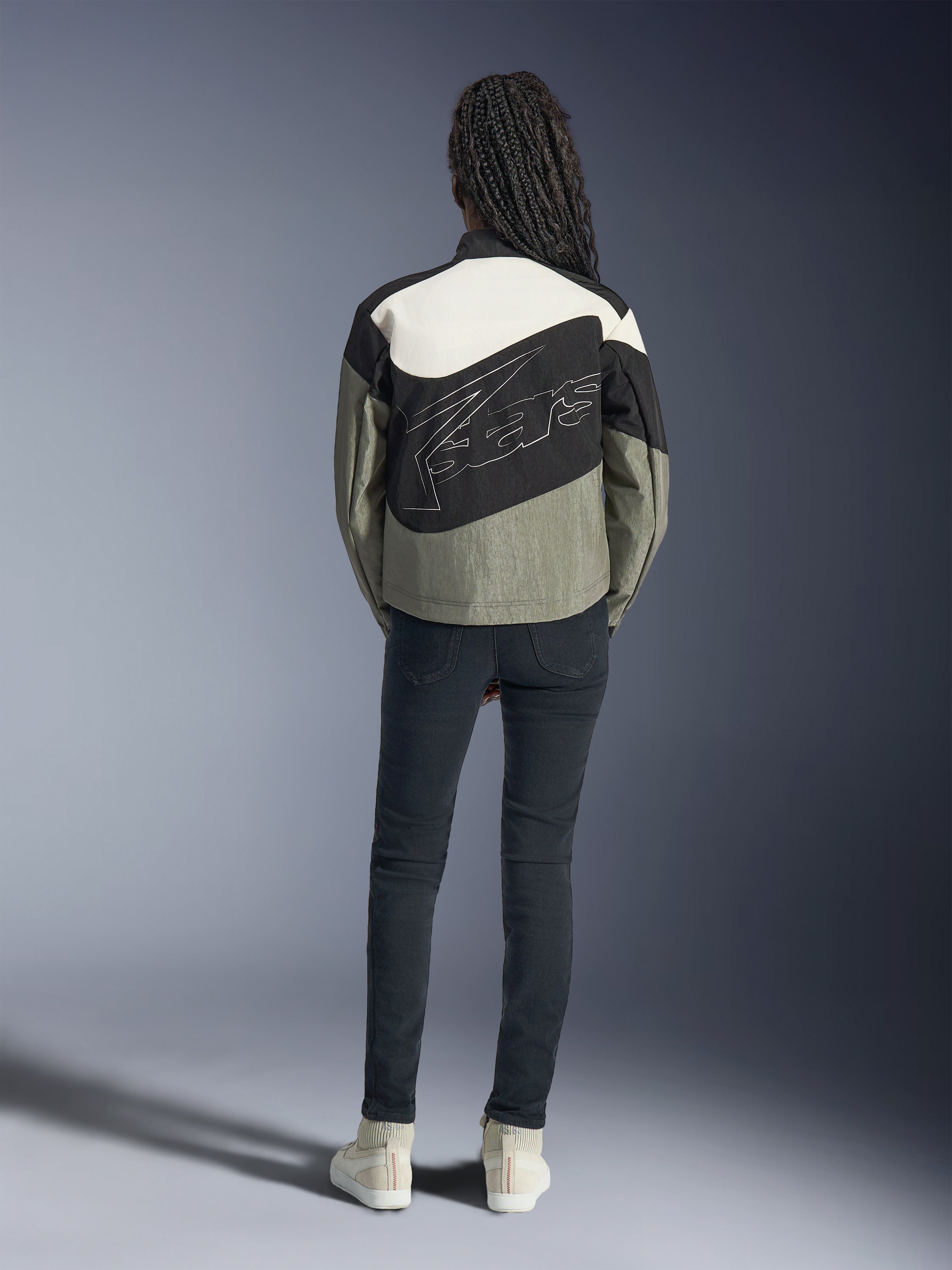 Rückansicht eines Models in der Alpinestars Stella Moshell Jacke in Schwarz, Grau und Off-White, mit asymmetrischem Design und großem, stilisiertem Logo auf dem Rücken, kombiniert mit einer dunklen Slim-Fit-Jeans und weißen Sneakern, urbane Streetwear-inspirierte Motorradbekleidung für Damen.