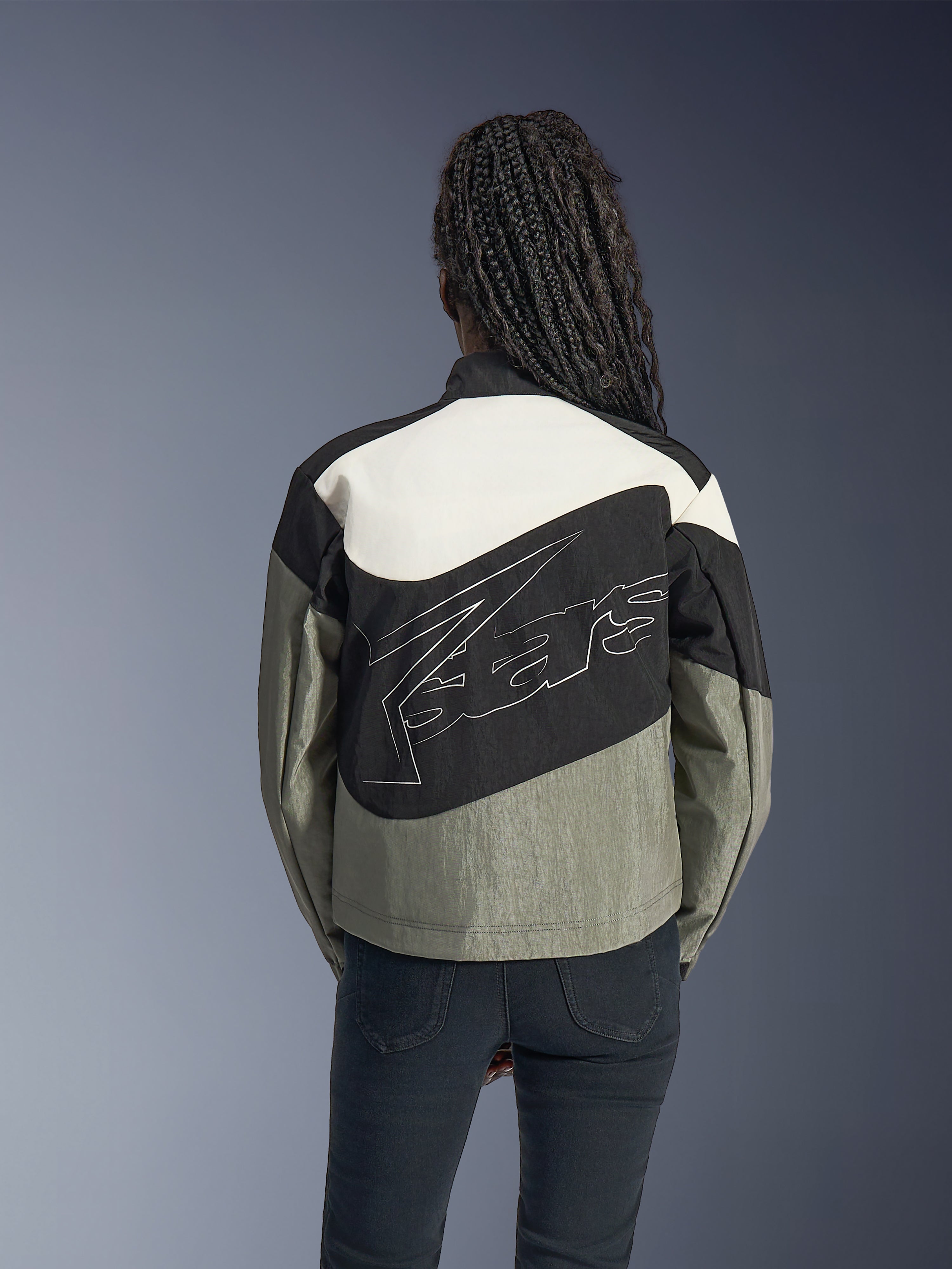Frau von hinten in der Alpinestars Stella Moshell Jacke, urbane Motorradjacke in Schwarz, Grau und Off-White, mit großem weiß umrandetem Logo auf dem Rücken, kombiniert mit schwarzer Hose, moderne von Streetwear inspirierte Motorradausrüstung.