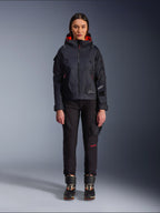 Alpinestars Actea Women's Pants, black motorcycle pants with red text detail and cargo pockets, worn by a female rider with a black hooded jacket, modern urban motorcycle apparel