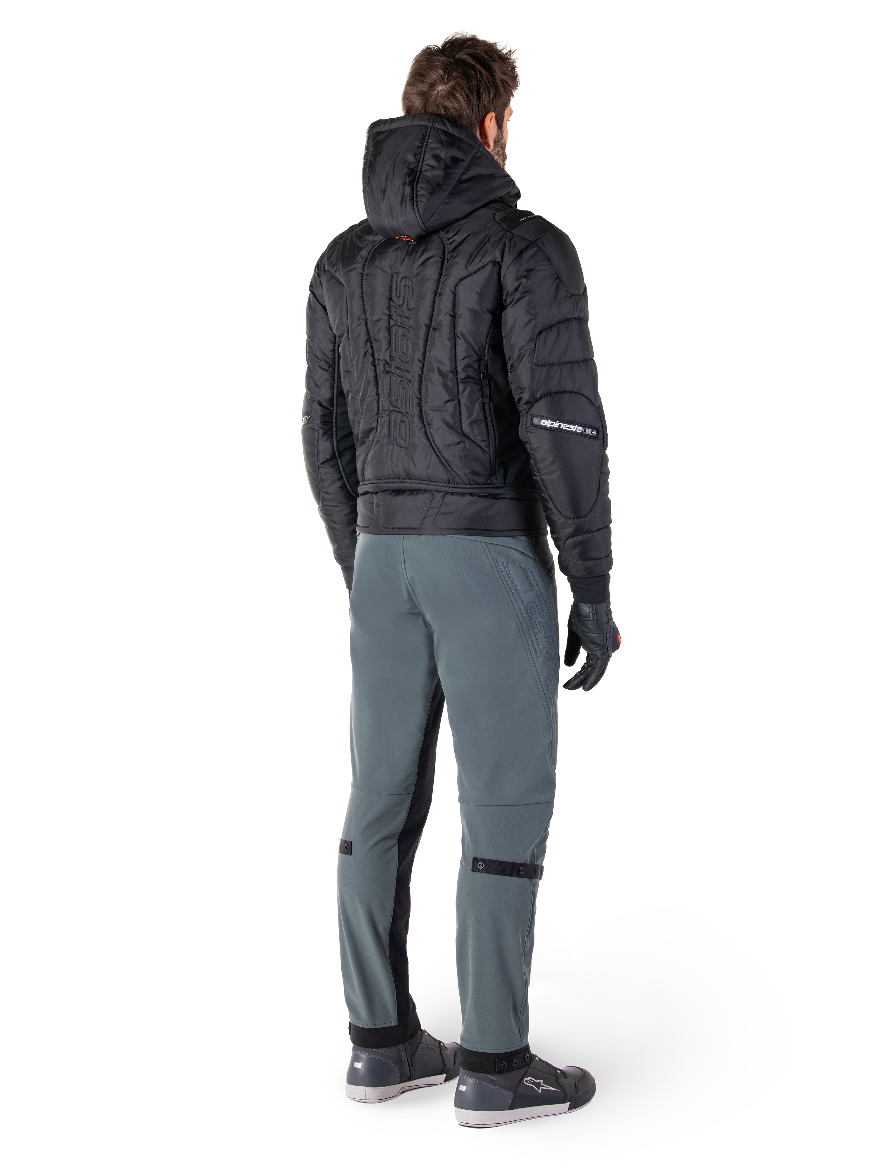 Alpinestars Alden Hose, urbane Motorradhose, Urban Gray, Rückansicht eines Fahrers in schwarzer gesteppter Kapuzenjacke und Motorrad-Sneakern, schmale Passform unterhalb des Knies mit schwarzen Stretcheinsätzen und Knieverstellern, modernes, von Streetwear inspiriertes Design.