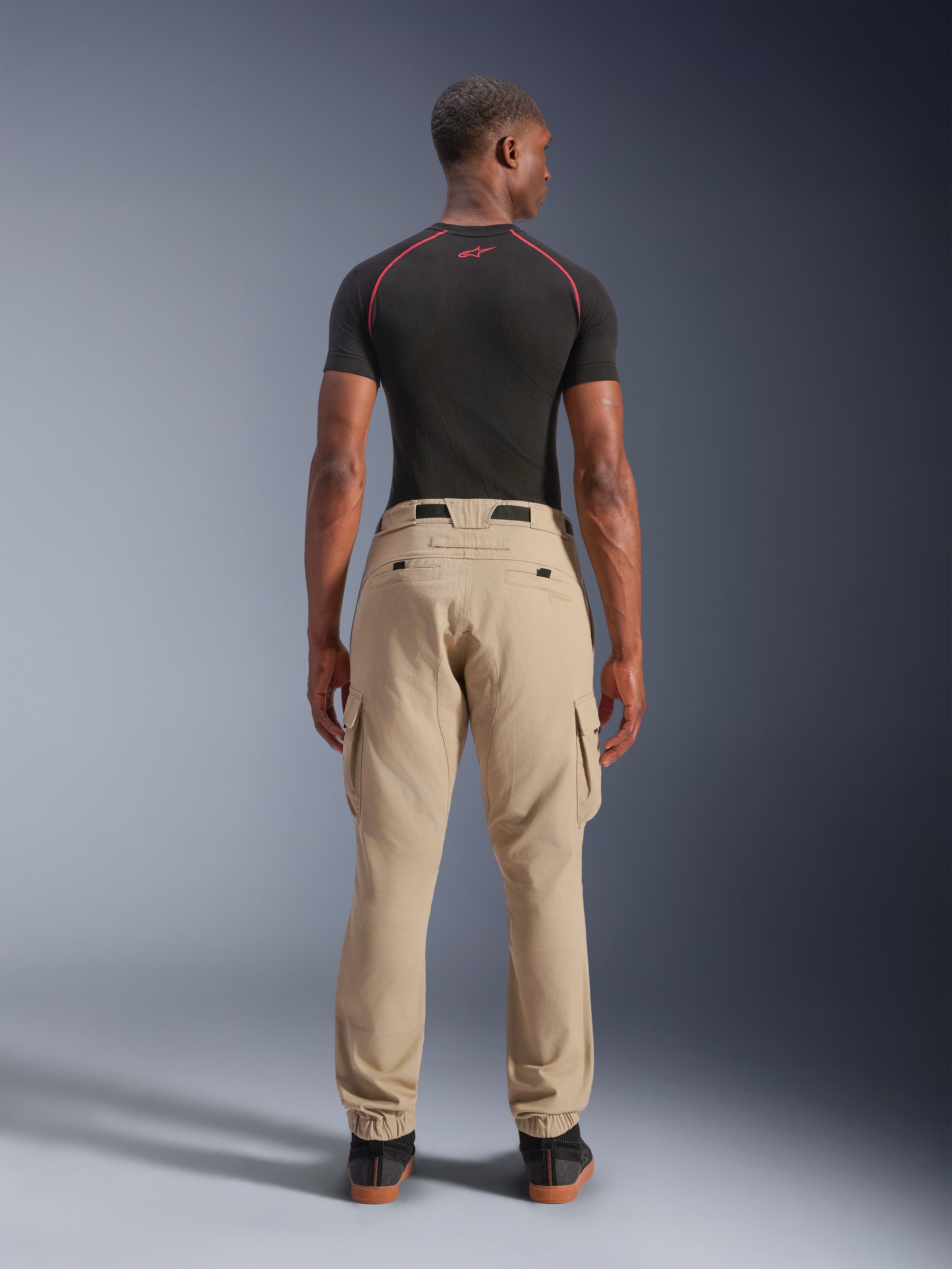 Alpinestars Flex-AST Cargo-Hose – Kurz, Hellbeige, Rückansicht an einem Model, reguläre Tapered-Passform mit elastischen Säumen und Cargotaschen, getragen mit einem schwarzen technischen Base Layer mit roten Akzenten, moderne urbane Motorradbekleidung