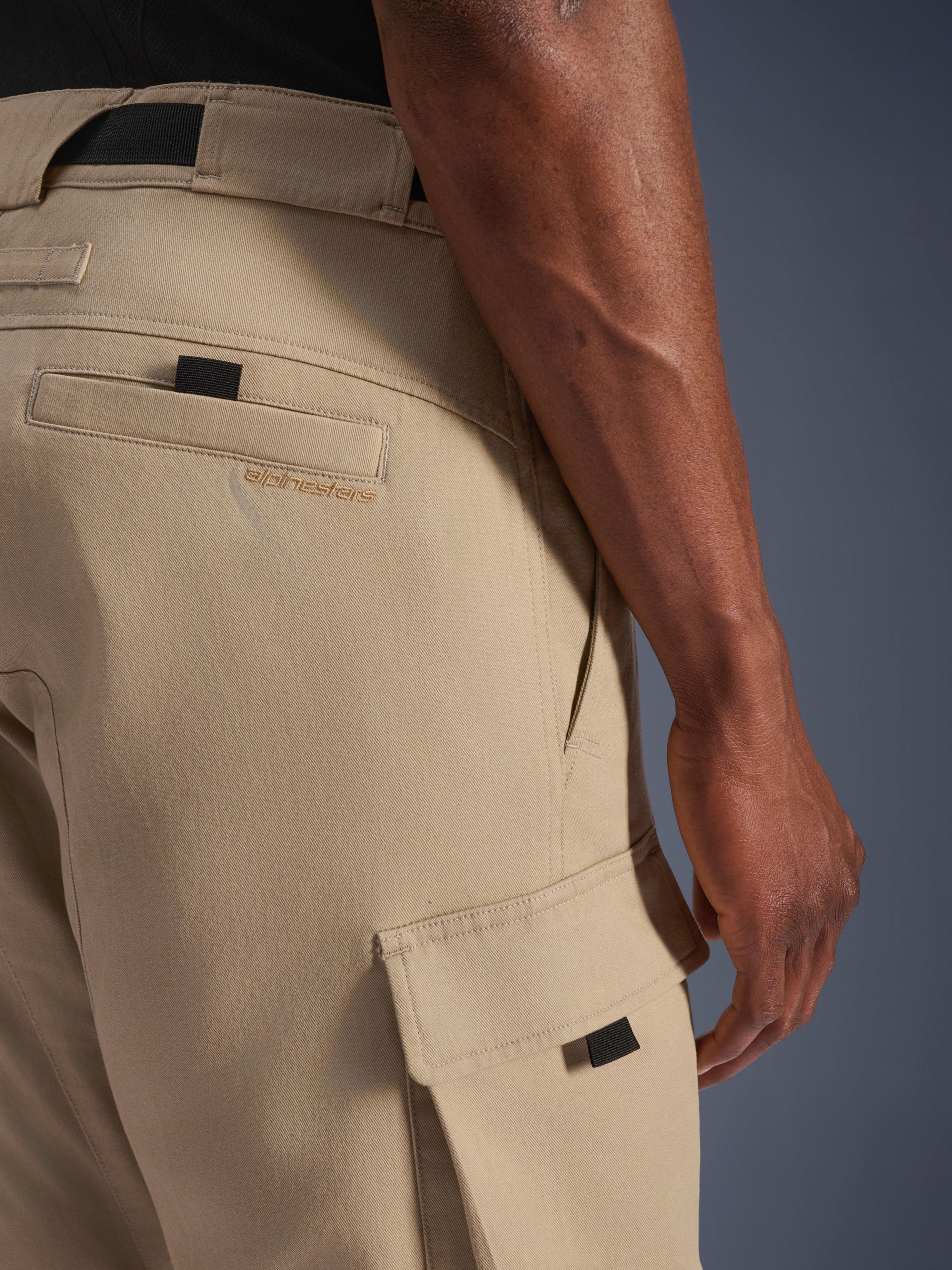 Alpinestars Flex-AST Cargo-Hose – Kurz, Hellbeige, Nahaufnahme der Seitenansicht mit Cargotasche samt schwarzer Zuglasche und gesticktem Logo auf der Gesäßtasche, strapazierfähiges Textilmaterial, urbane Motorradausrüstung.