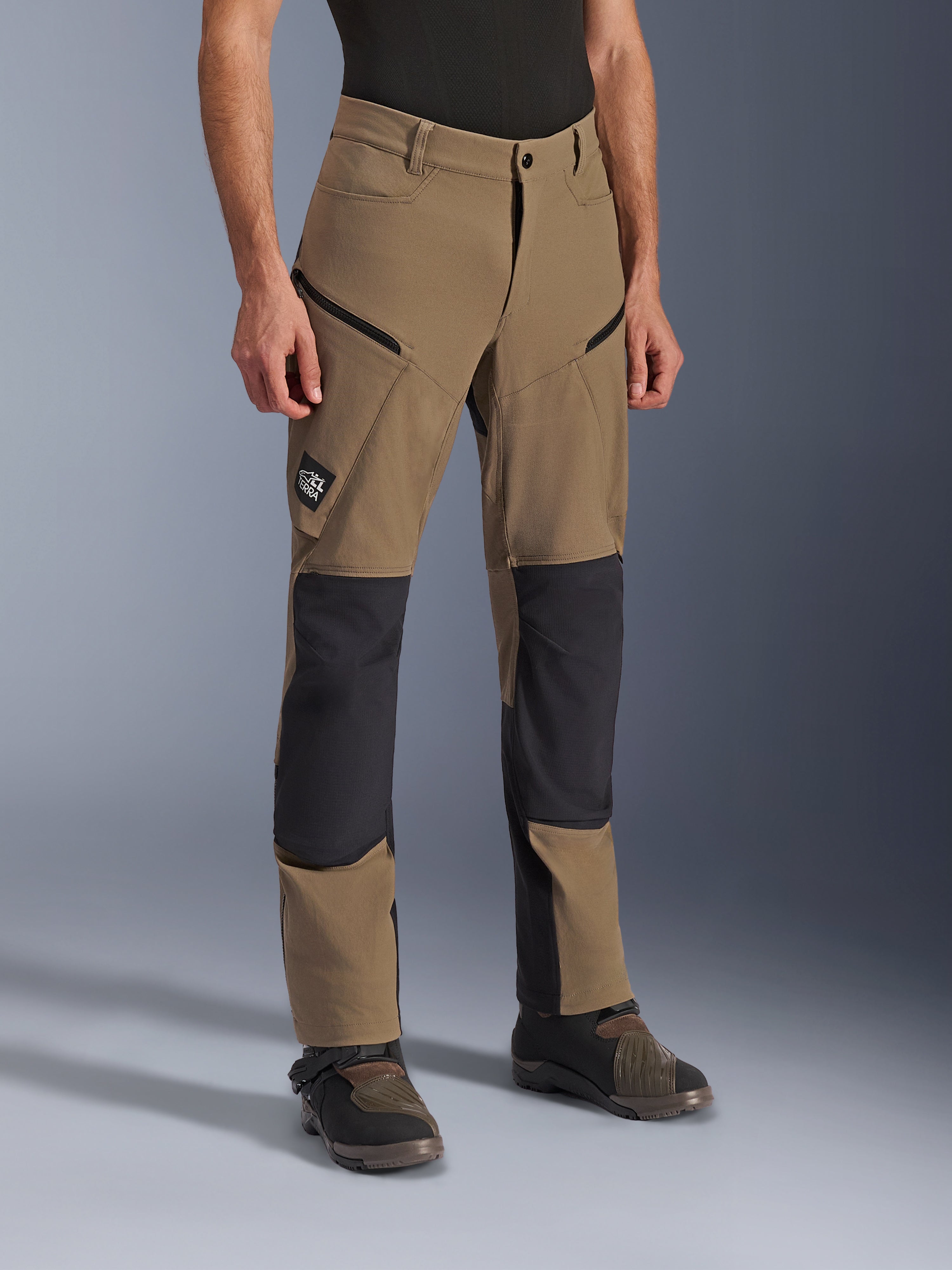 Alpinestars Flex-AST Explorer Pants, adventure motorcycle pants in Walnut Black, featuring tan textile with black reinforced knee and seat panels, zippered cargo pockets, and All Terra logo patch, shown worn with technical riding boots.
