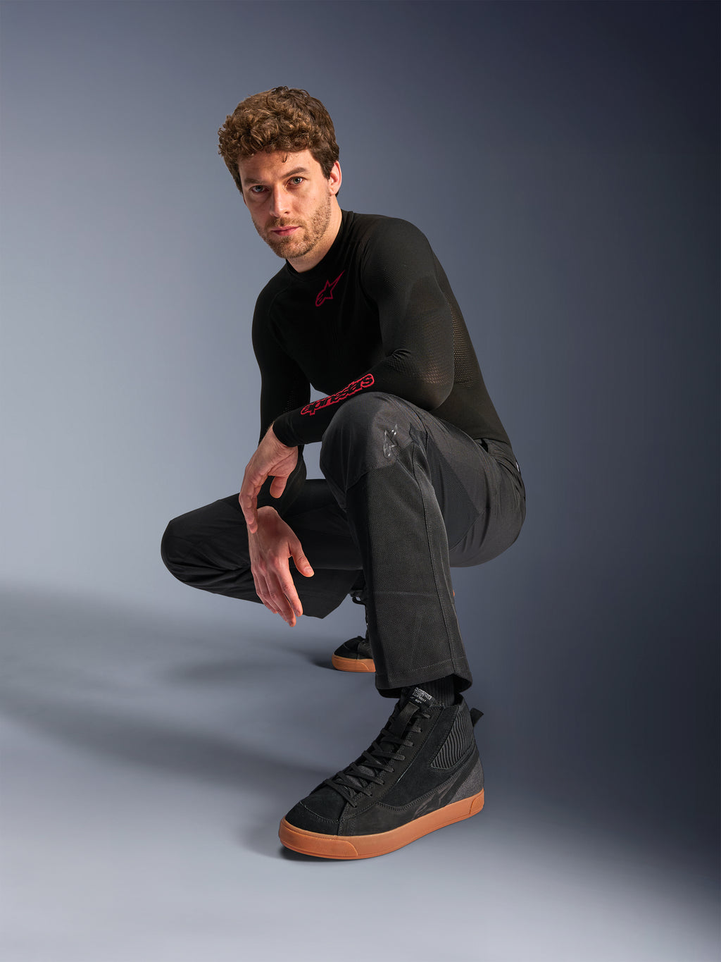 Alpinestars C-1 Air Hose, Urbane Motorradhose, Schwarz, Modell hockend in einer Studio-Umgebung, kombiniert mit schwarzem Langarm-Oberteil und schwarzen Sneakern mit hellbraunen Sohlen, mit atmungsaktiven Mesh-Einsätzen und modernem urbanen Stil