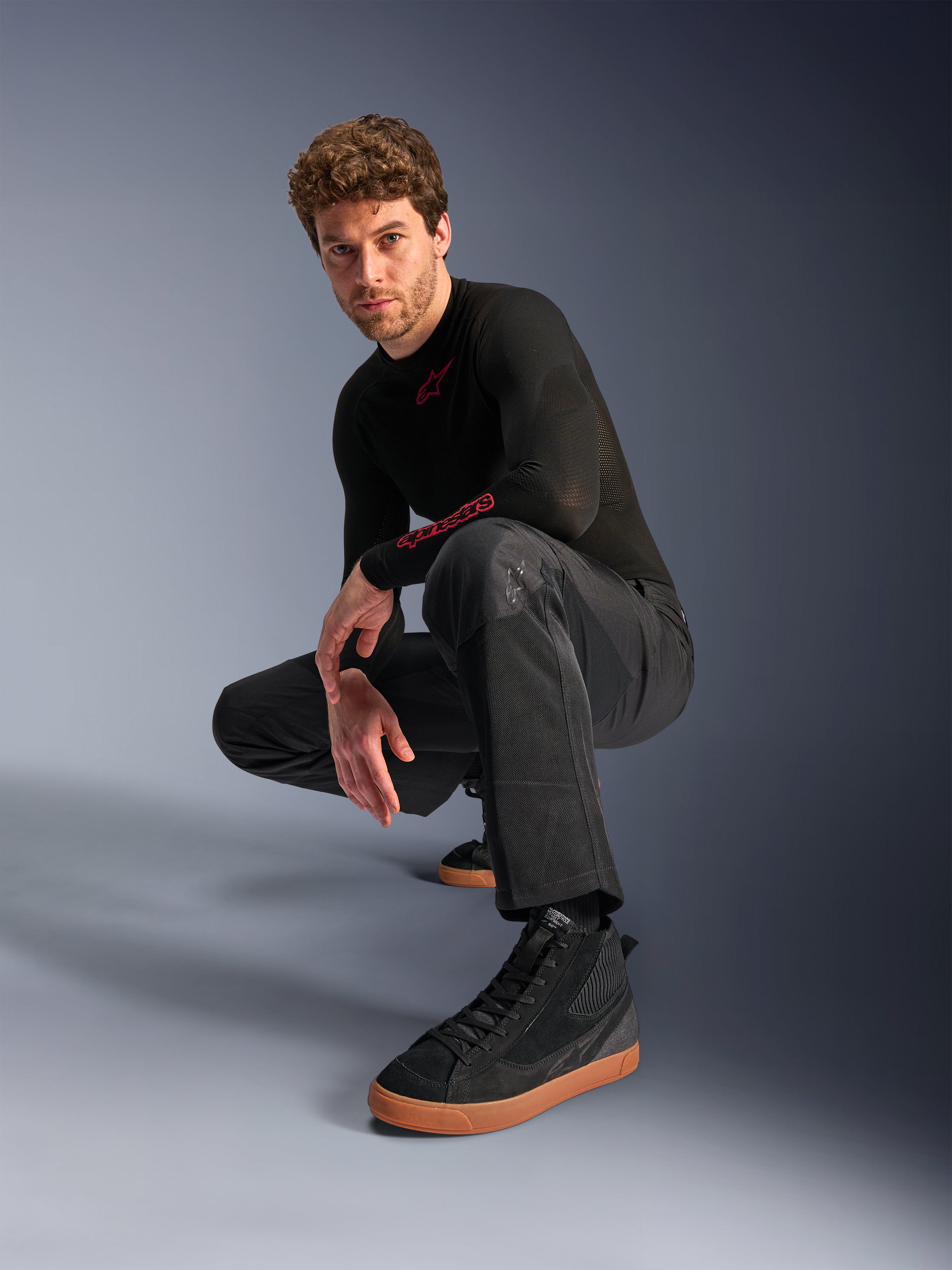 Alpinestars C-1 Air Hose, Urbane Motorradhose, Schwarz, Modell hockend in einer Studio-Umgebung, kombiniert mit schwarzem Langarm-Oberteil und schwarzen Sneakern mit hellbraunen Sohlen, mit atmungsaktiven Mesh-Einsätzen und modernem urbanen Stil