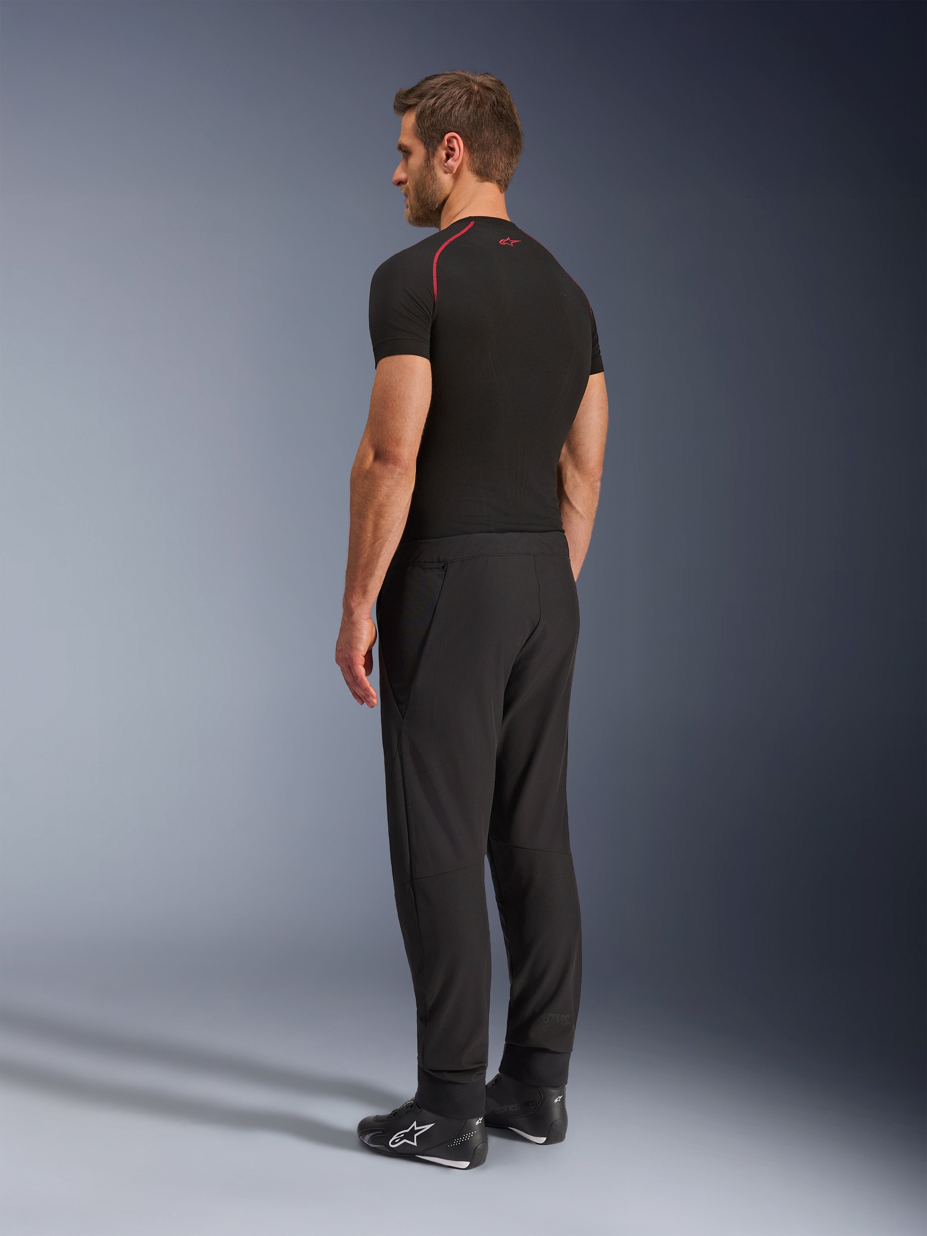 Alpinestars Aeroshell Airflow Pants, Black motorcycle pants, lightweight softshell textile, worn with a black technical short-sleeve shirt and black riding boots, rear-side profile view showing the slim fit and ergonomic construction.