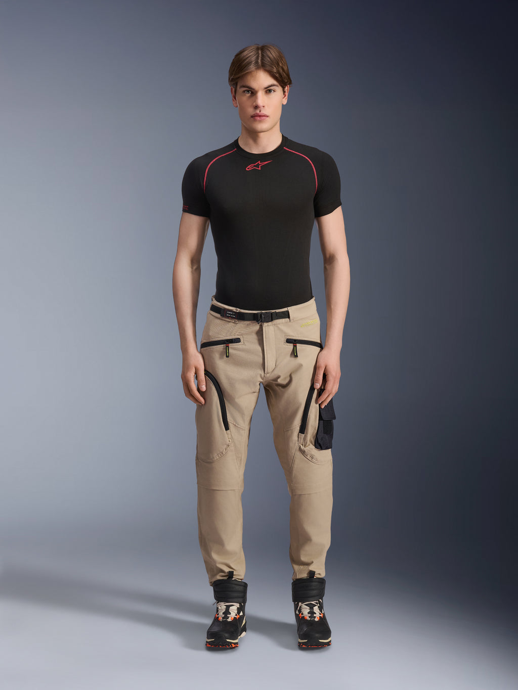 Männliches Model in Alpinestars Acteon Hose in Sand, kombiniert mit einem schwarzen kurzärmeligen Base Layer und schwarzen technischen Stiefeln, mit Cargotaschen mit schwarzen Reißverschlüssen und einer schmal zulaufenden Passform, zeitgemäße urbane Motorradausrüstung