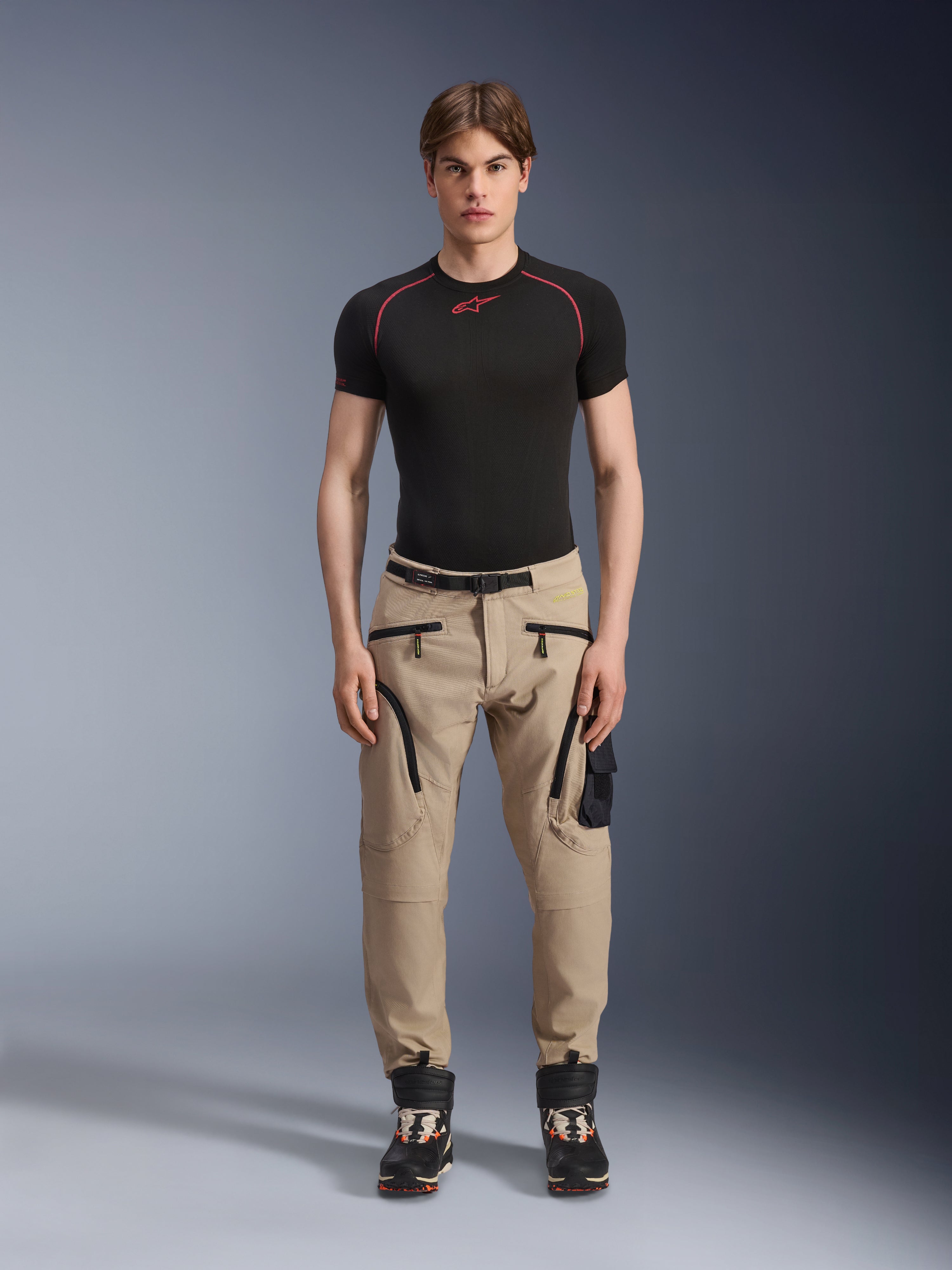 Männliches Model in Alpinestars Acteon Hose in Sand, kombiniert mit einem schwarzen kurzärmeligen Base Layer und schwarzen technischen Stiefeln, mit Cargotaschen mit schwarzen Reißverschlüssen und einer schmal zulaufenden Passform, zeitgemäße urbane Motorradausrüstung