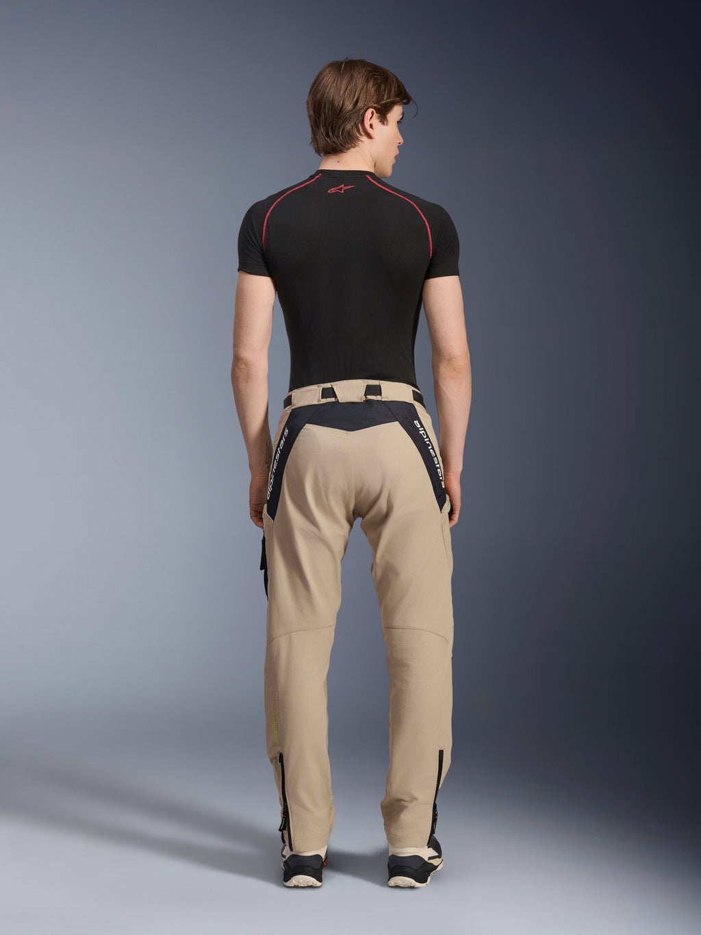 Alpinestars Acteon Hose in Sand, moderne urbane Motorradhose, Rückansicht, schwarze Einsätze mit weißem Alpinestars-Branding, schmal zulaufende Passform mit Reißverschlüssen am Saum, getragen mit schwarzem kurzärmeligem Baselayer und Sneakern.